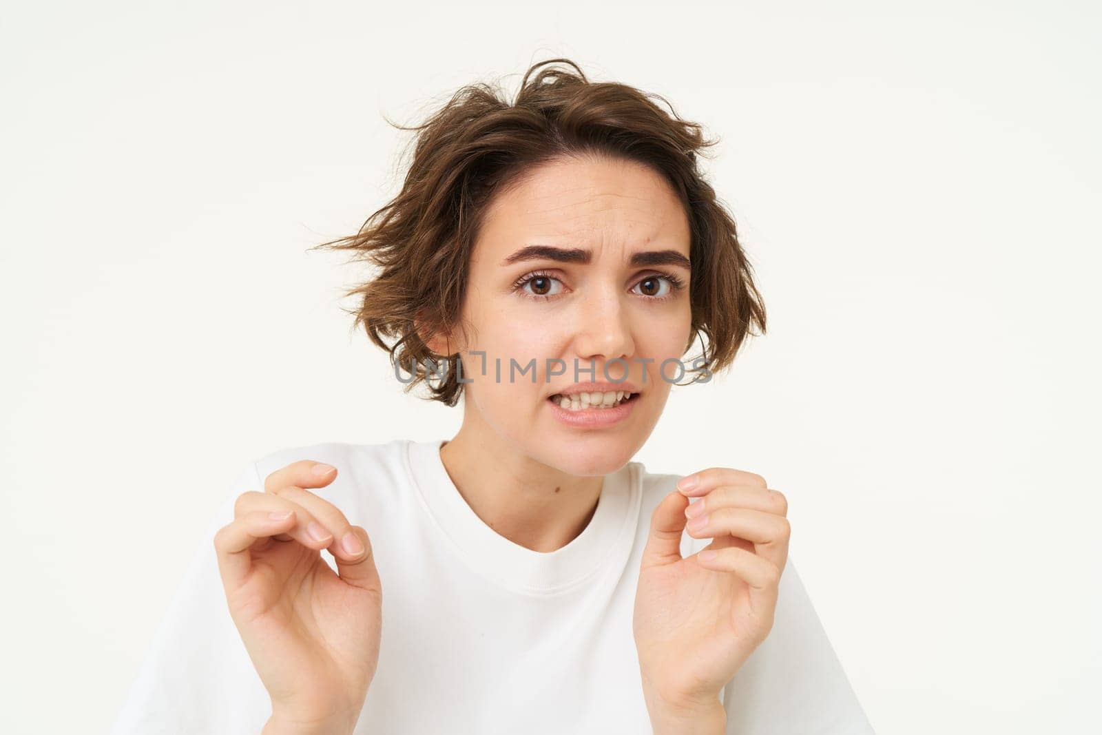 Portrait of woman jumps from fear, looks disgusted, cringe from something, express dislike or aversion, stands over white background.