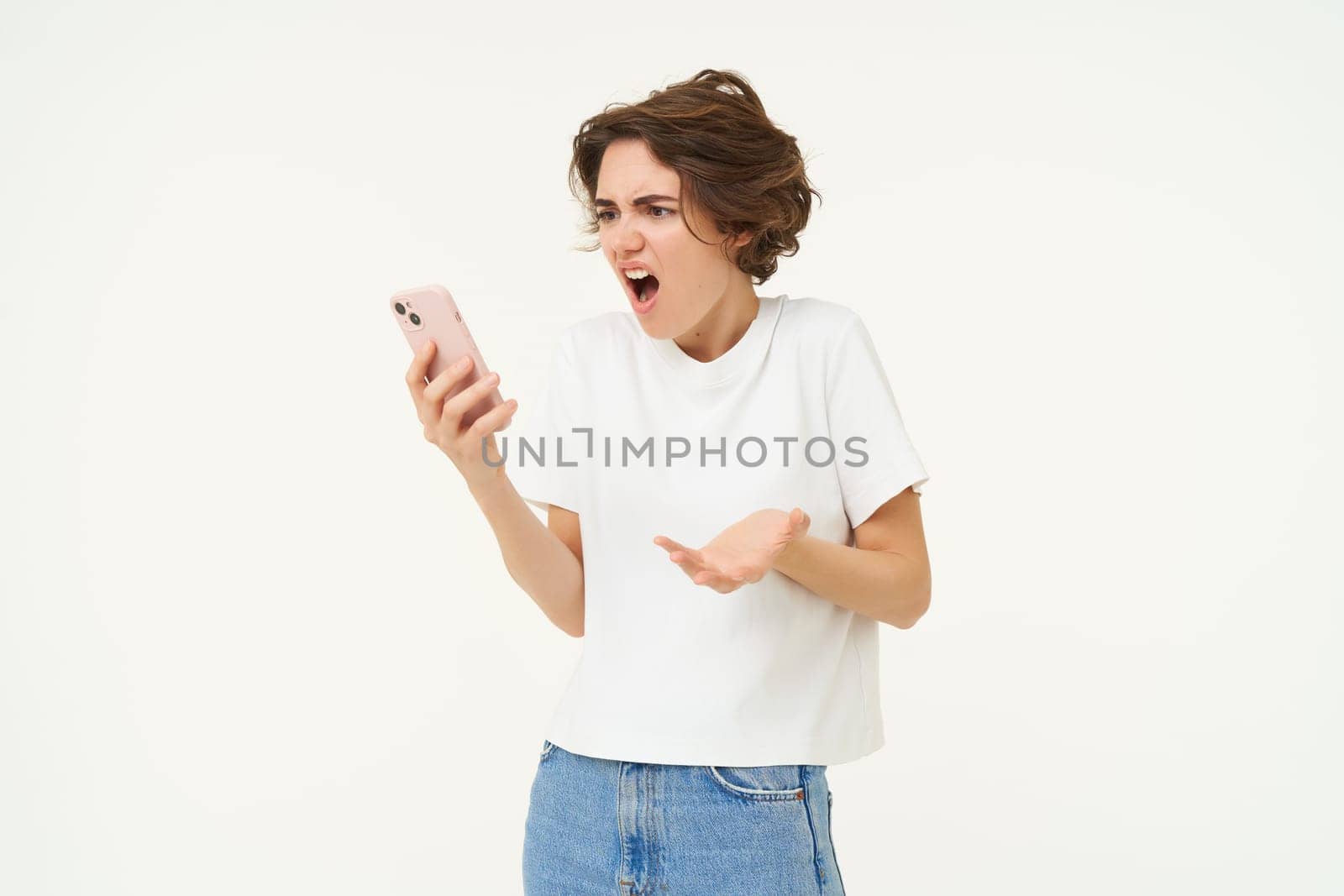 Portrait of confused and upset young woman, holding smartphone, shrugging shoulders and complaining at something online, standing over white background.