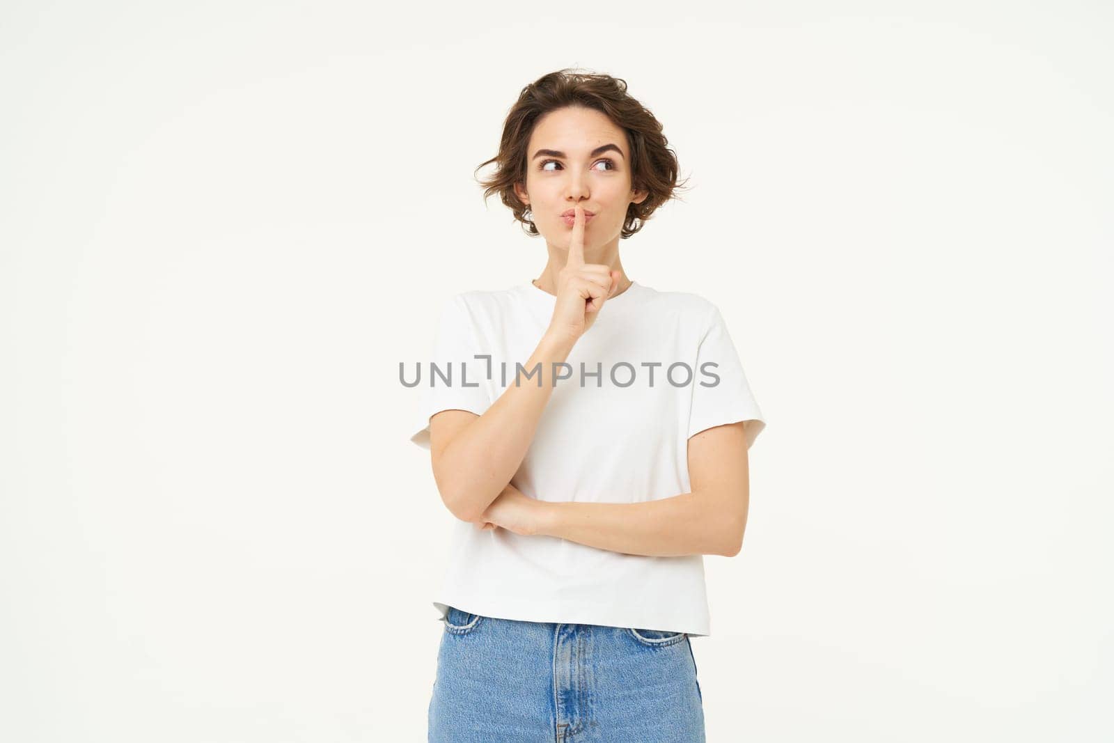 Image of brunette woman hiding secret, press finger to lips, hush, shush shh gesture, gossips, whispers, stands over white studio background.