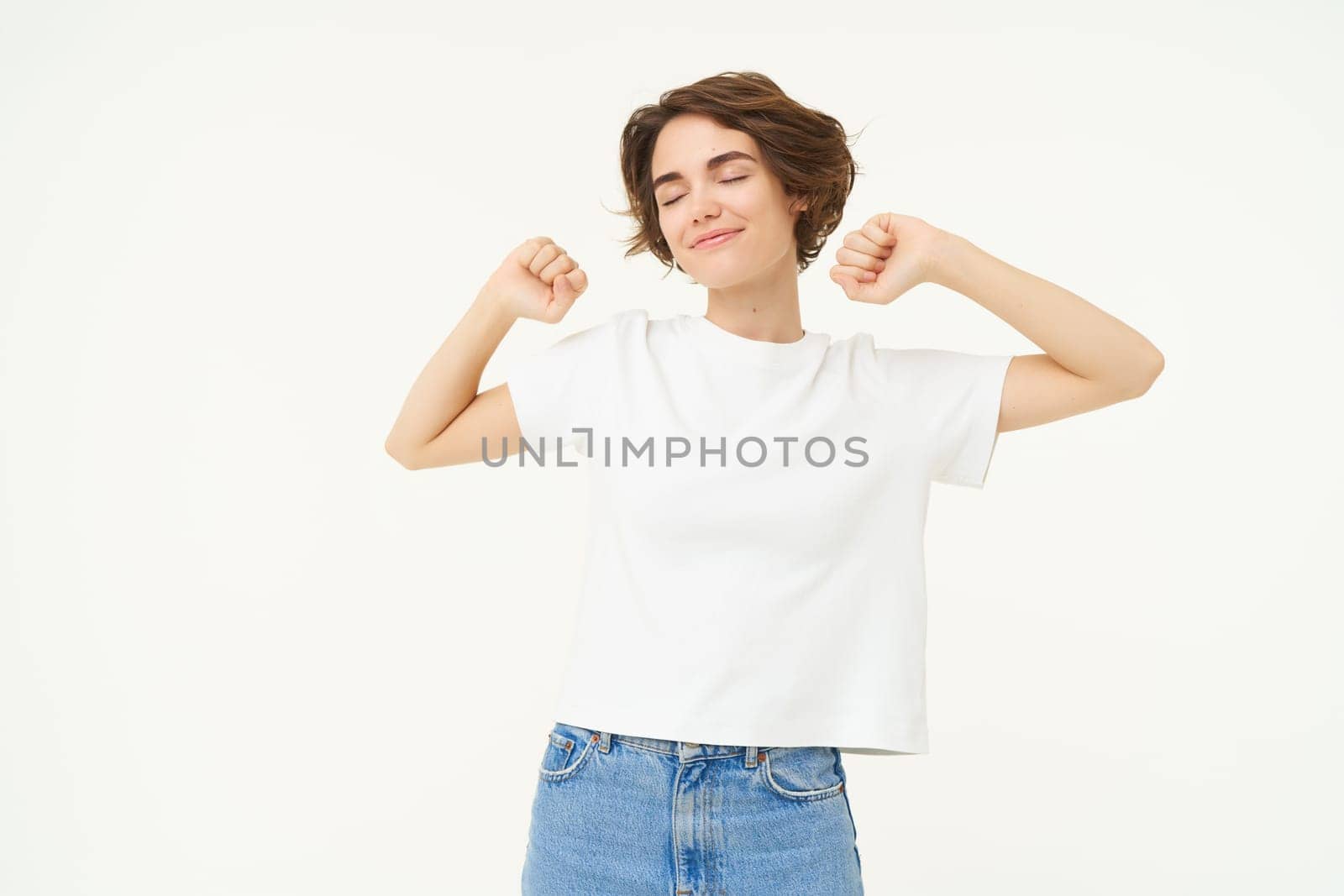 Portrait of smiling, pleased woman stretching hands, waking up after sleep, standing over white background by Benzoix