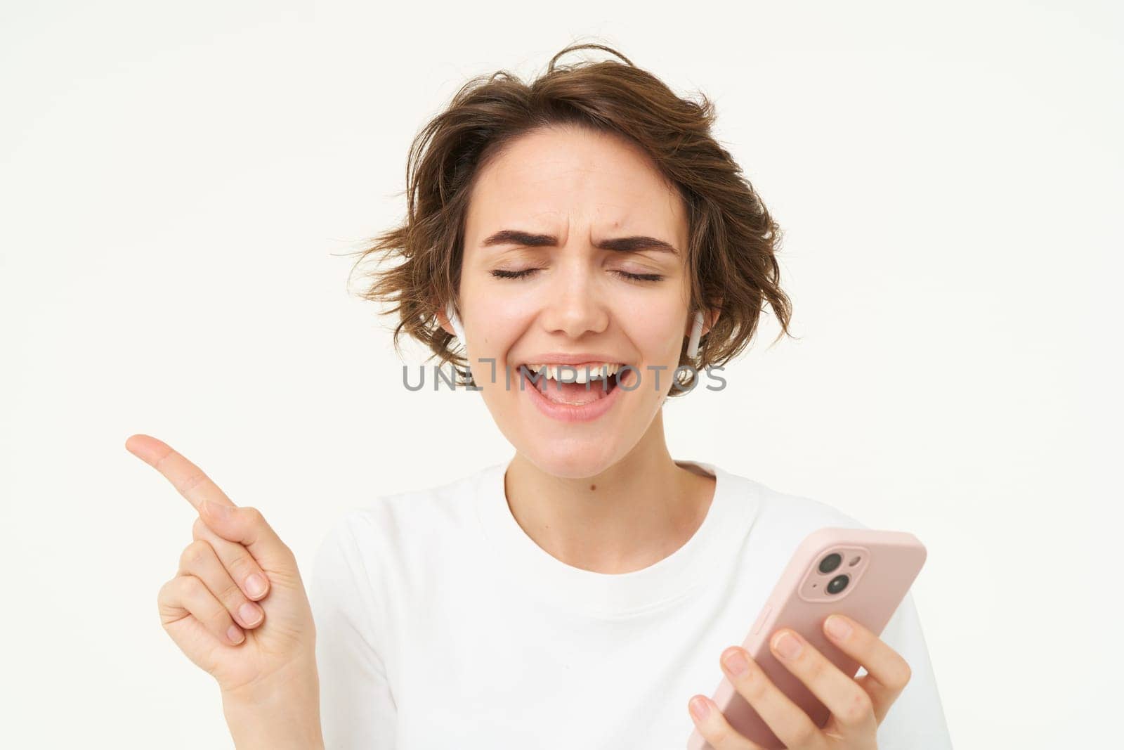 Woman enjoys listening to music in her earphones, holding smartphone, choosing song on streaming service app, standing over white background by Benzoix