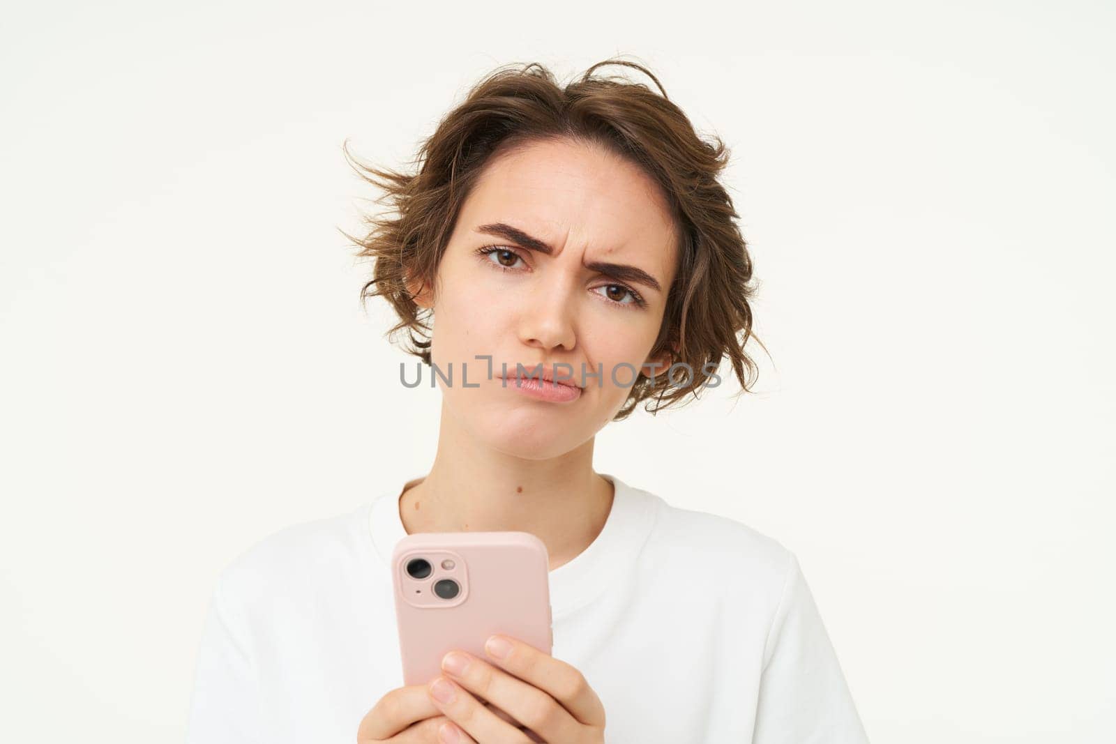 Image of complicated, confused young woman, frowning, holding smartphone, looking puzzled and doubtful, standing over white background.