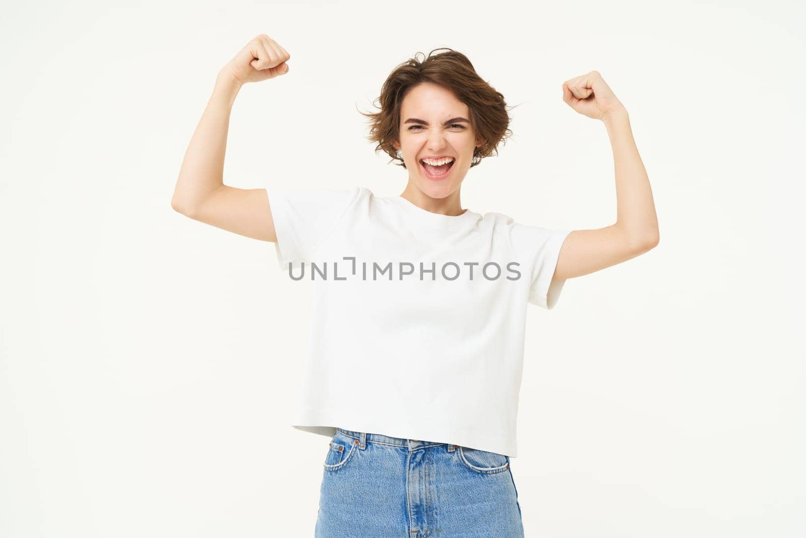 Portrait of happy and confident girl shows muscles, flexing biceps and smiling, posing over white background by Benzoix