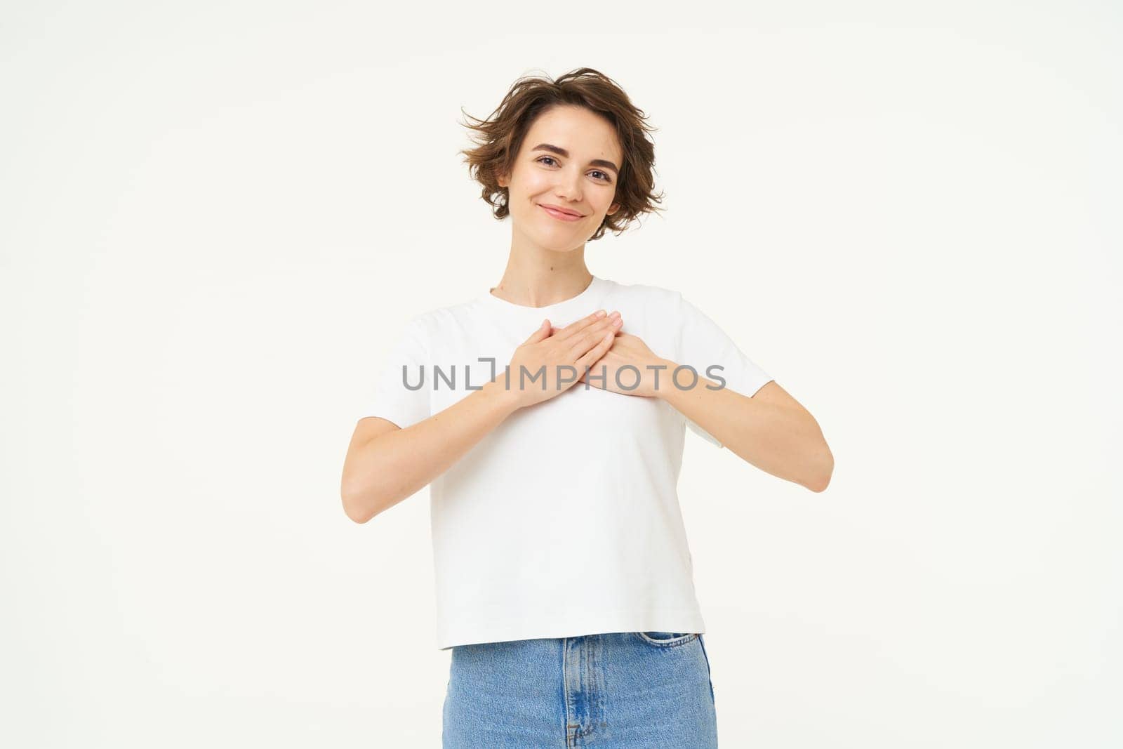 Portrait of brunette woman with hands on chest, looks with care and love, express gratitude, self-care and heart-warming feelings, stands over white background by Benzoix