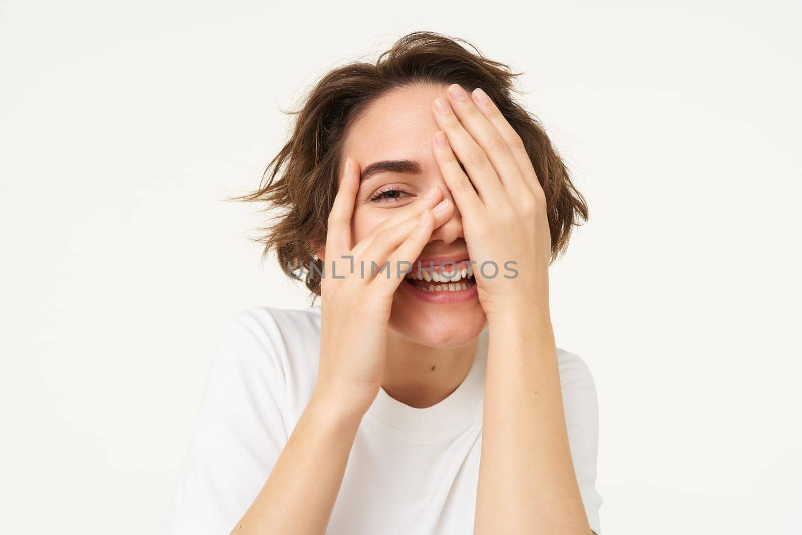 Genuine young woman, laughing and smiling, peeking through fingers and looking at surprise, standing over white background. Copy space
