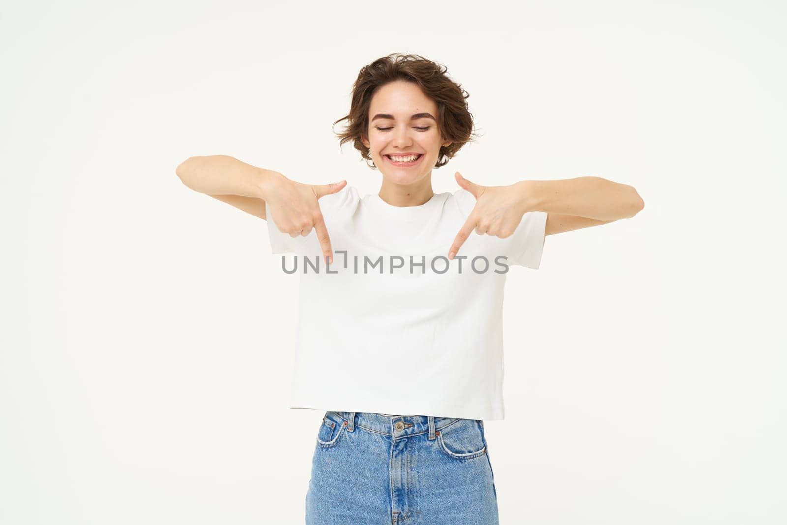 Image of beautiful, happy brunette woman, pointing fingers down, showing advertisement, standing over white studio background by Benzoix