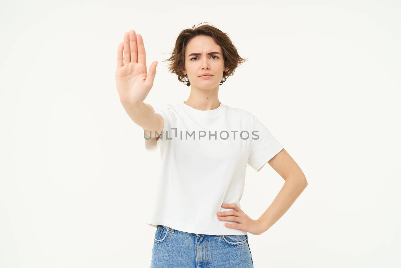 Portrait of serious, confident young woman extends one hand, shows stop, disapproval gesture, looking determined to block way, isolated over white background.