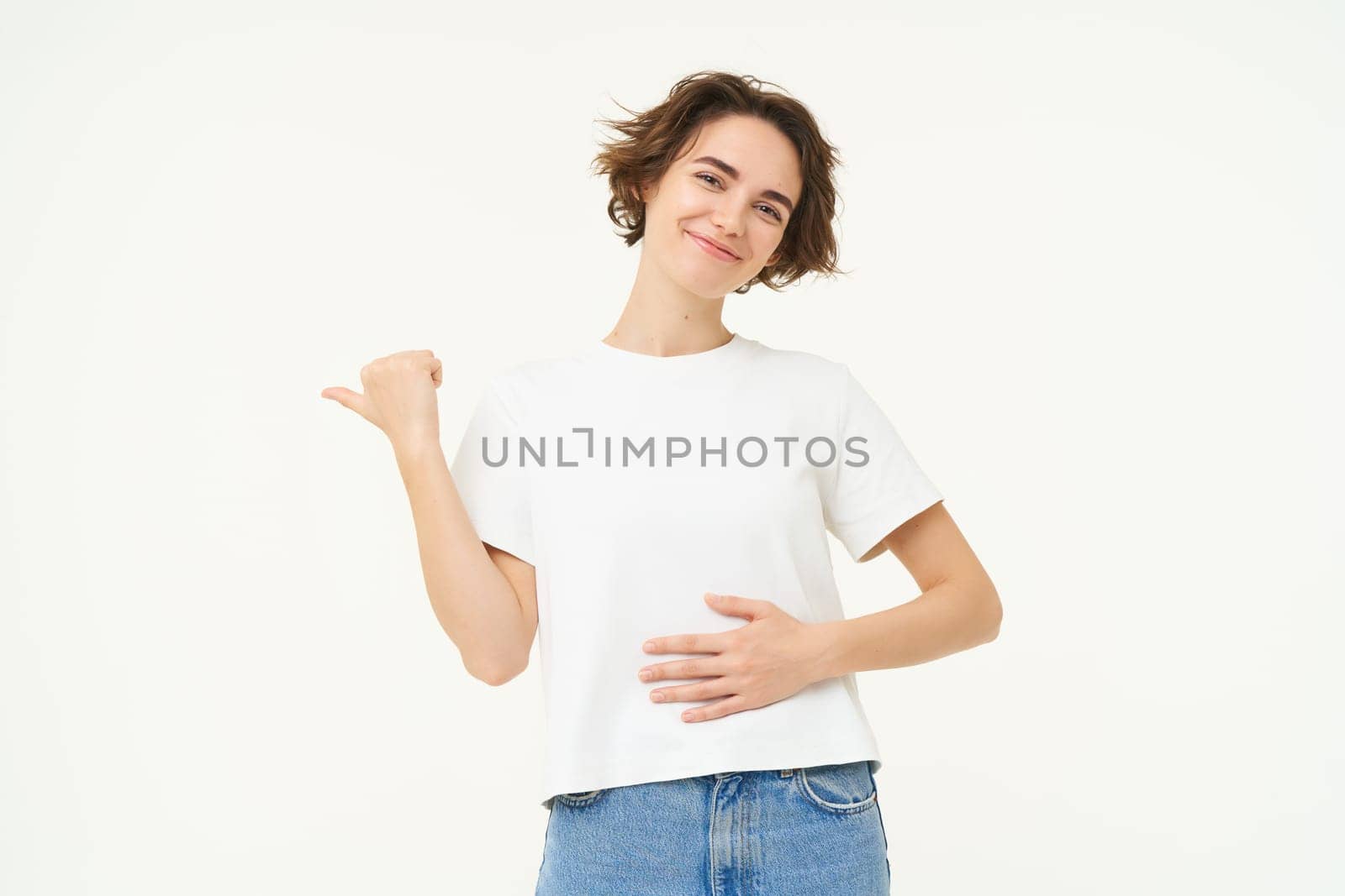 Portrait of smiling, pleased brunette woman, touches belly and points left, shows banner, recommends painkiller, pleased by medication effect on her stomach, stands over white background.