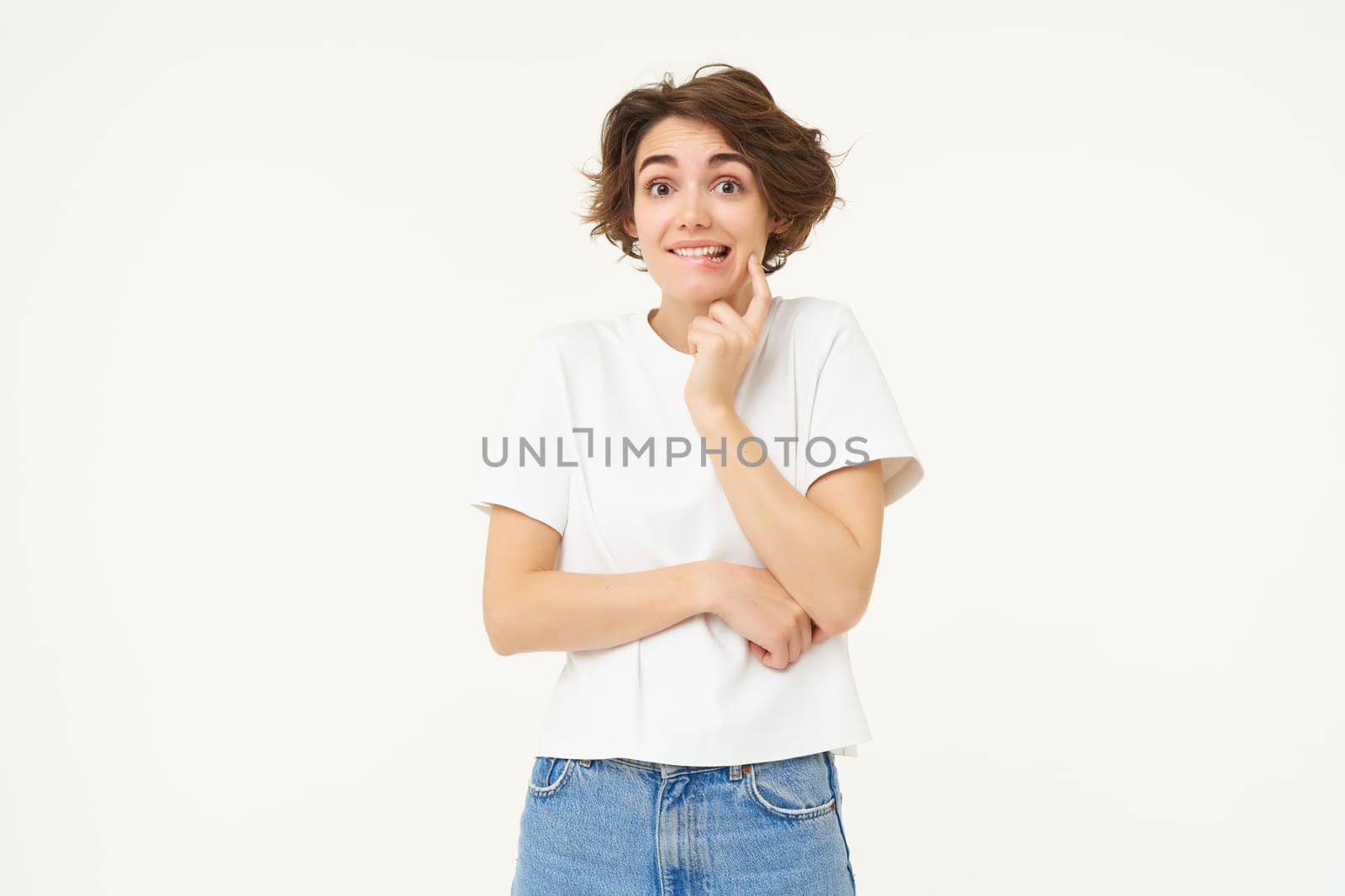 Portrait of cute and shy young woman, looking unsure at camera, posing in white t-shirt against studio background.