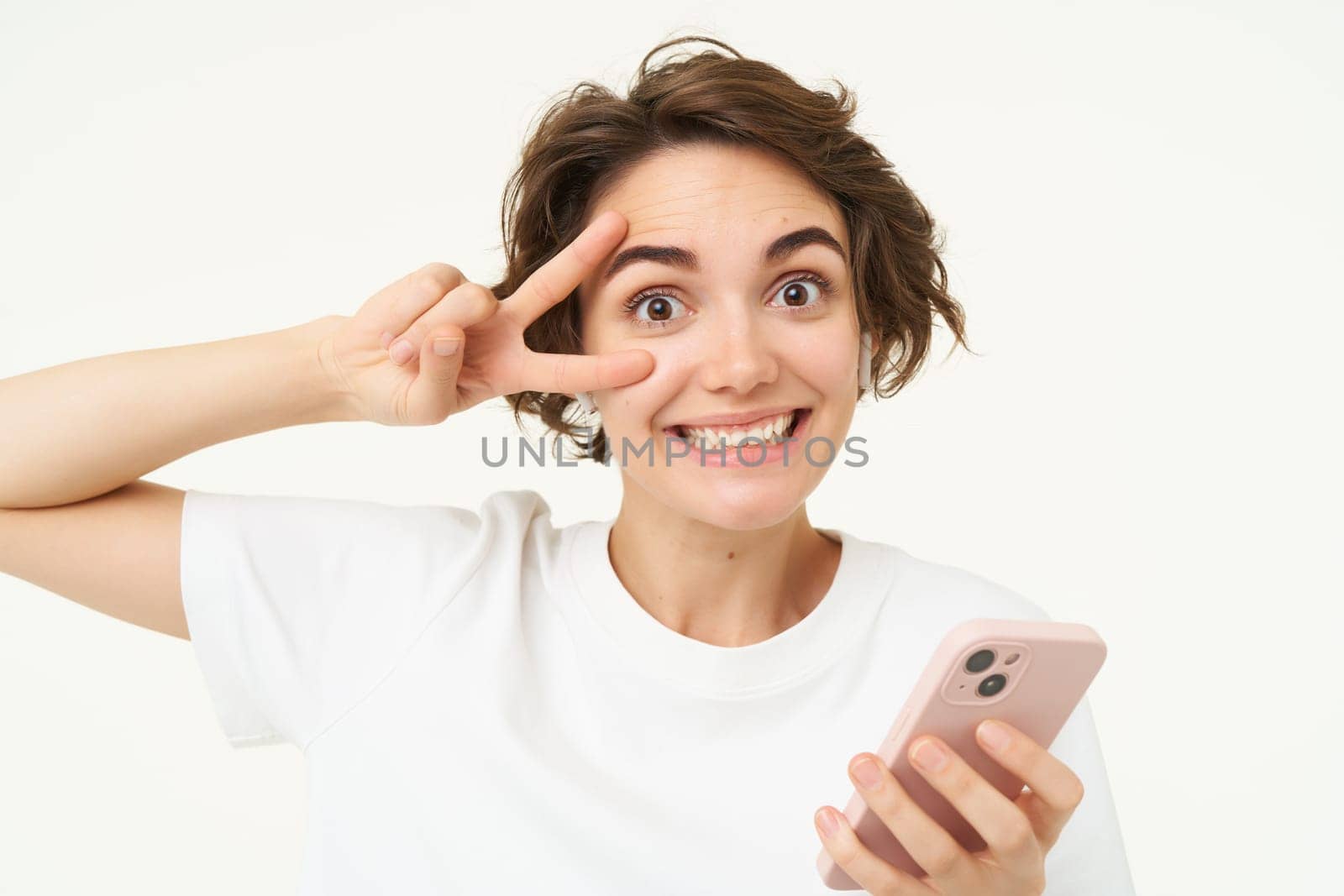 Portrait of young woman shows peace sign, holds smartphone, listens to music in wireless headphones, stands over white background.