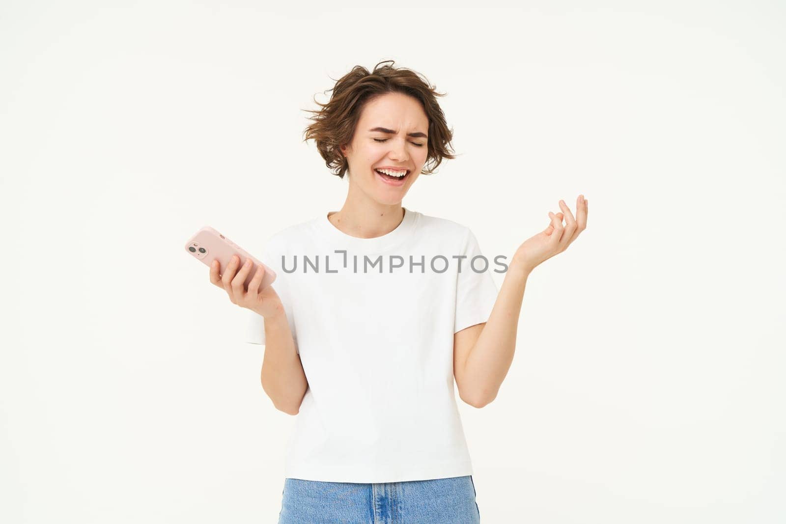 Portrait of brunette girl laughing, holding smartphone and giggle from something funny, posing over white studio background. Technology concept