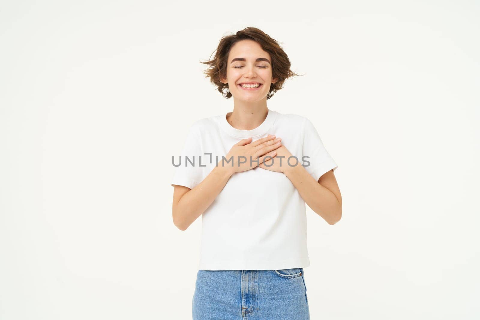 Image of brunette woman holding hands on heart, express gratitude, heartwarming feelings, emotion of happiness, love and care, isolated over white background.