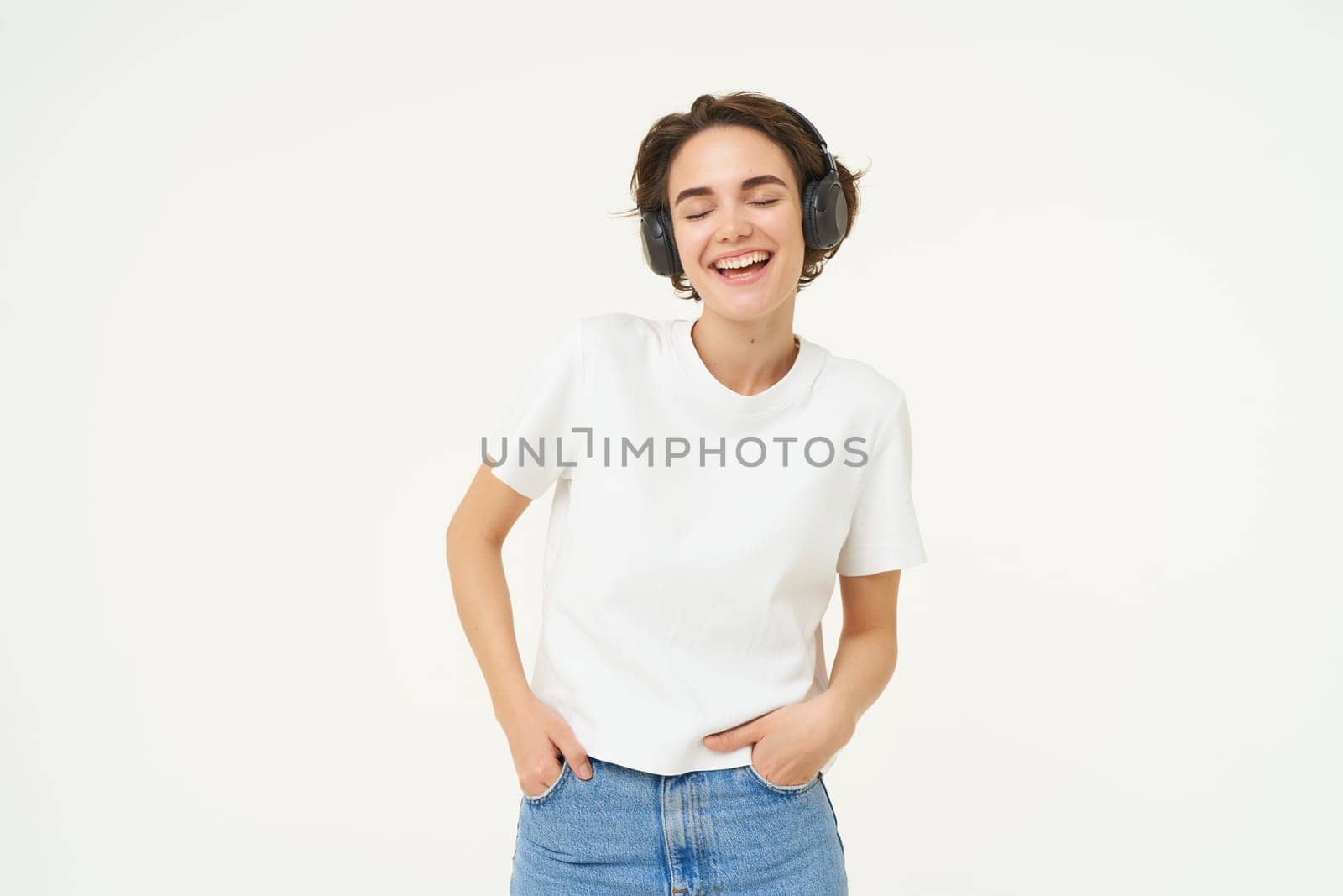 Portrait of young female model, wearing headphones, listens to music, smiling and laughing, standing over white background.