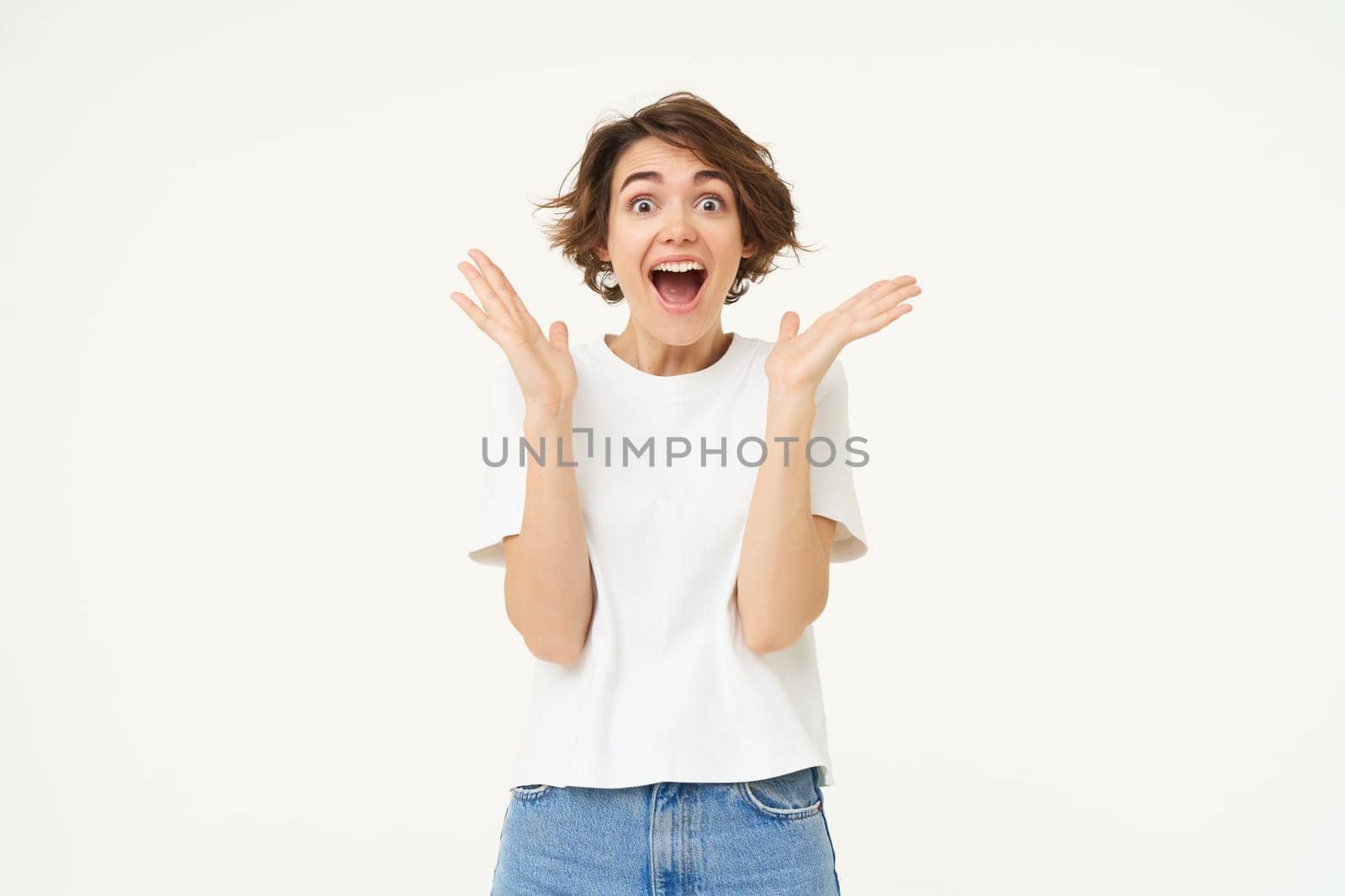 Image of woman with surprised face, amazed, says wow, claps hands and looks excited, stands over white background.