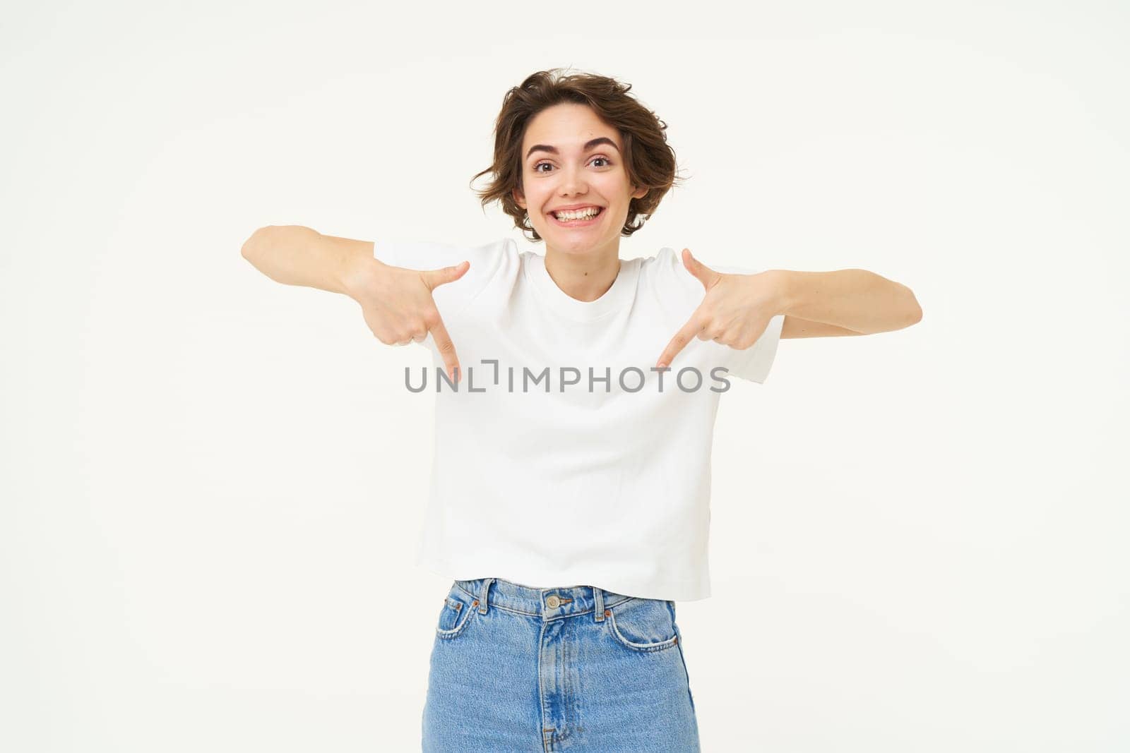 Portrait of cute, happy young woman pointing fingers down, showing advertisement, demonstrating banner on the bottom, standing against white background.