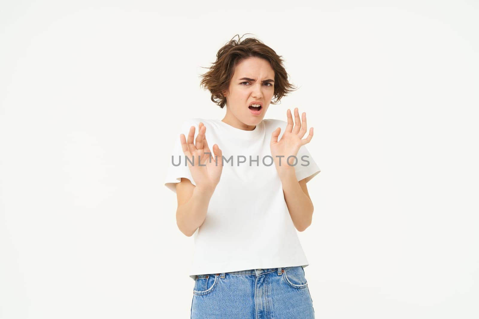 Young woman raises hands up and refuses, rejects smth, tried to block something disgusting, standing over white background.