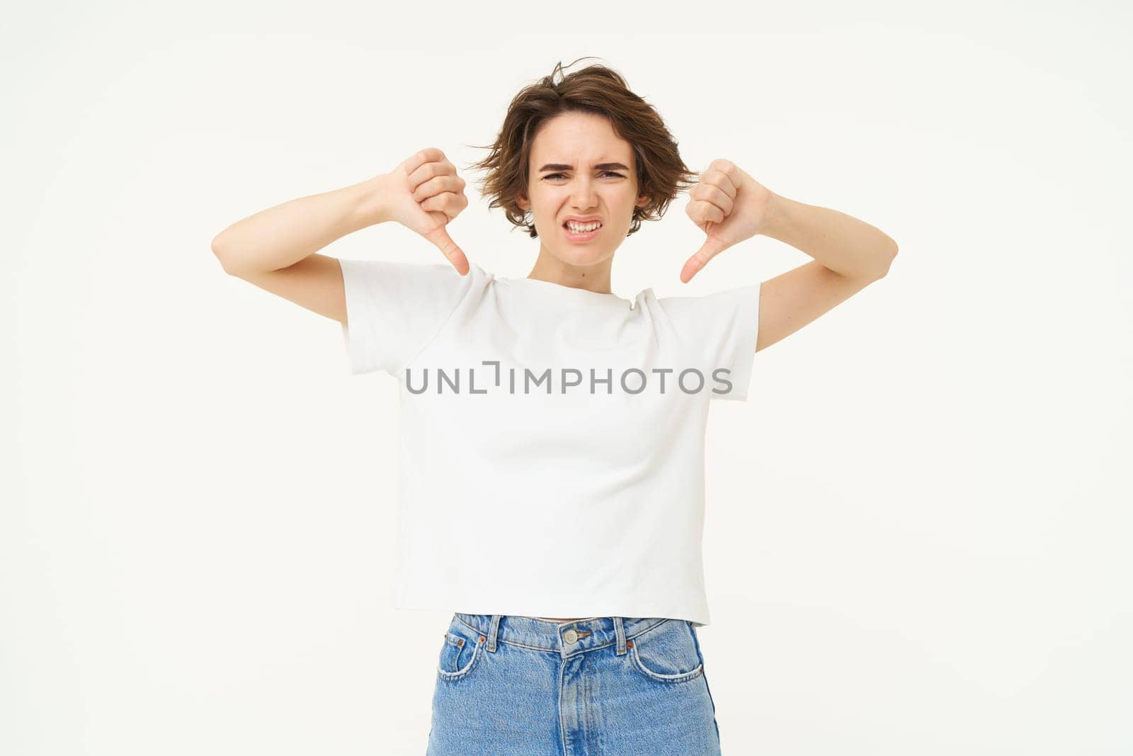 Portrait of disappointed young woman, student shows thumbs down from dislike, disapprove something bad, standing over white background.