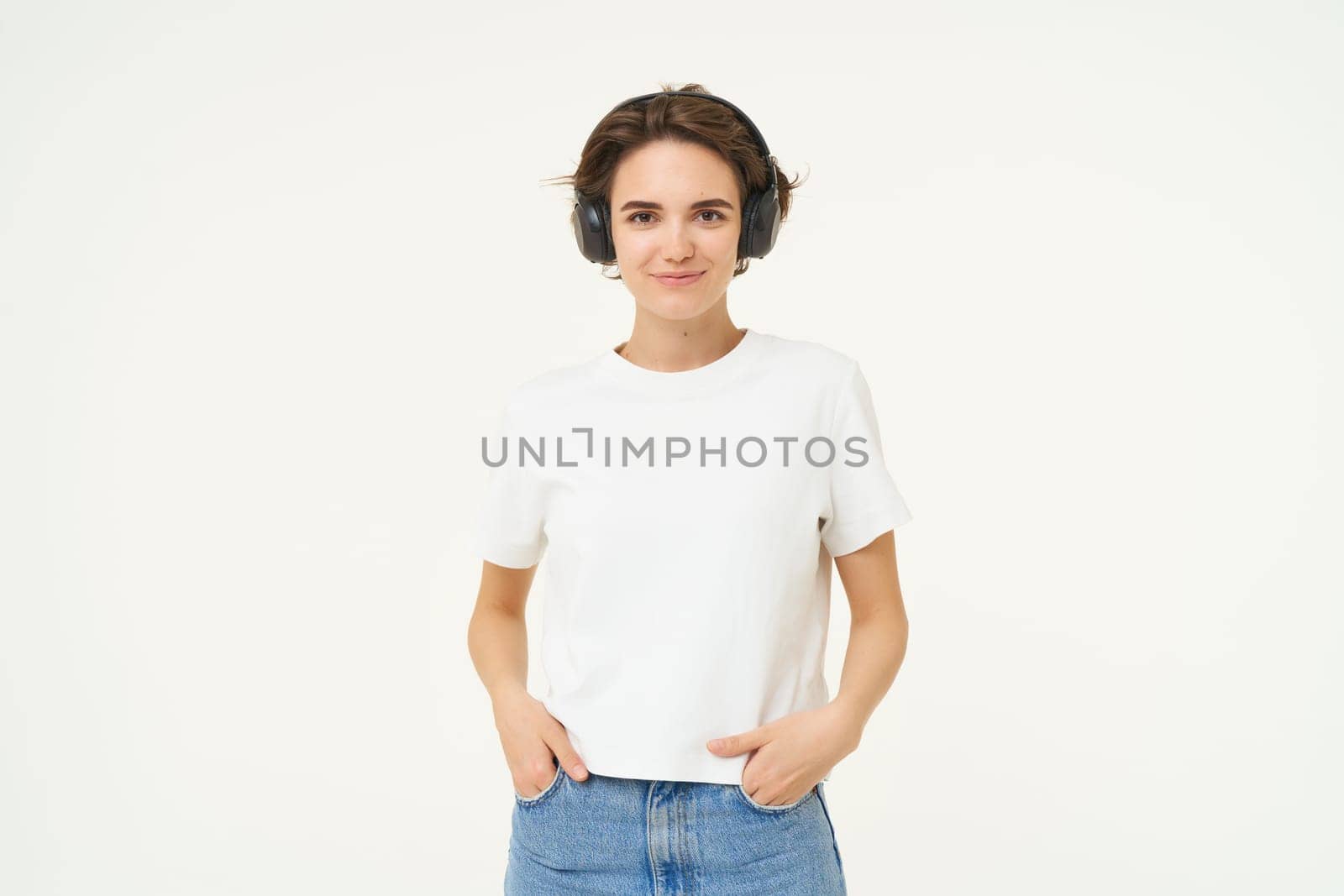 Portrait of young female model, wearing headphones, listens to music, smiling and laughing, standing over white background.