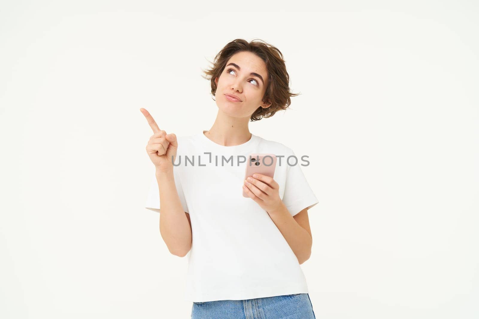 Portrait of young modern woman with mobile phone, looking up and pointing at advertisement, standing against white studio background.