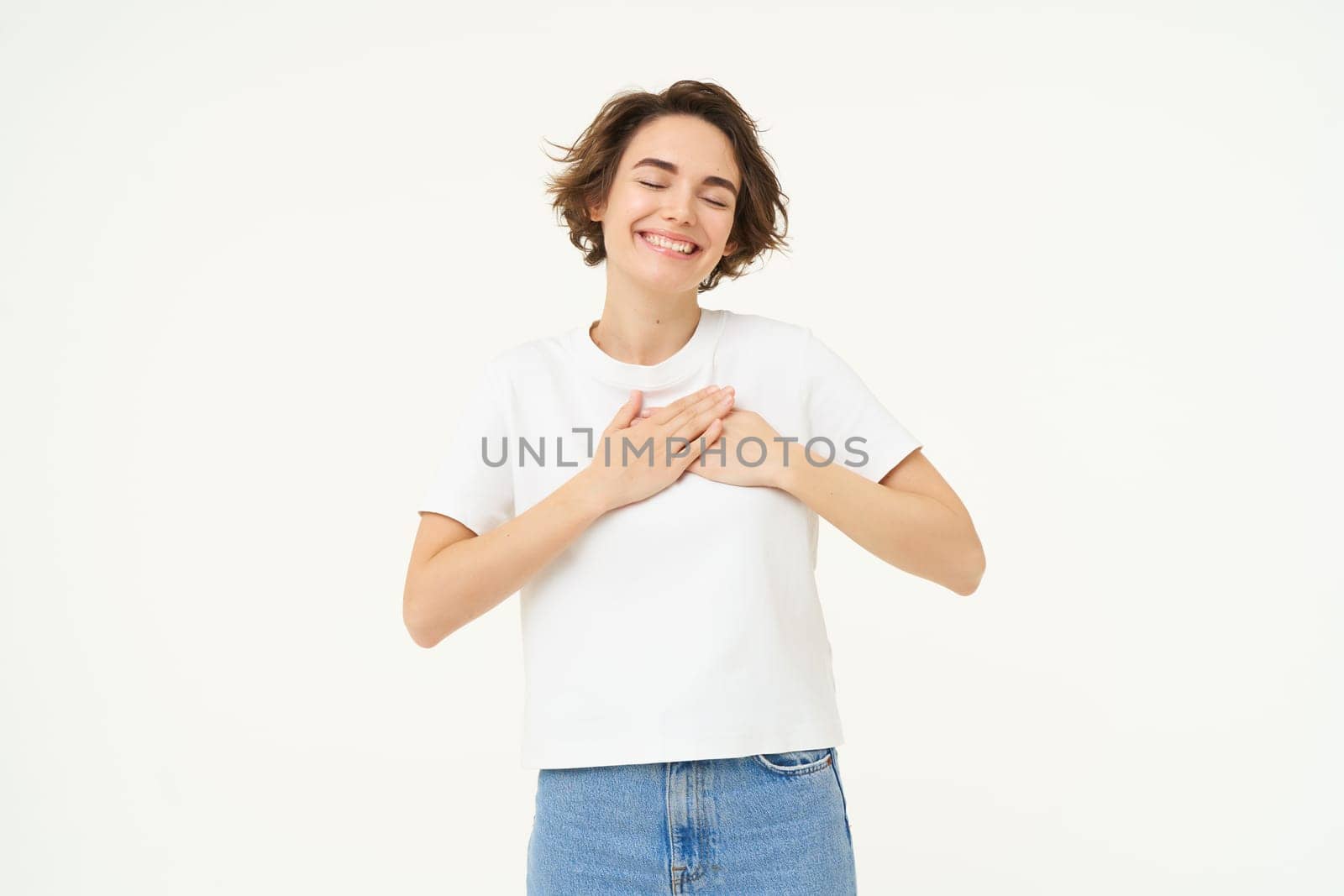 Image of brunette woman holding hands on heart, express gratitude, heart-warming feelings, emotion of happiness, love and care, isolated over white background.