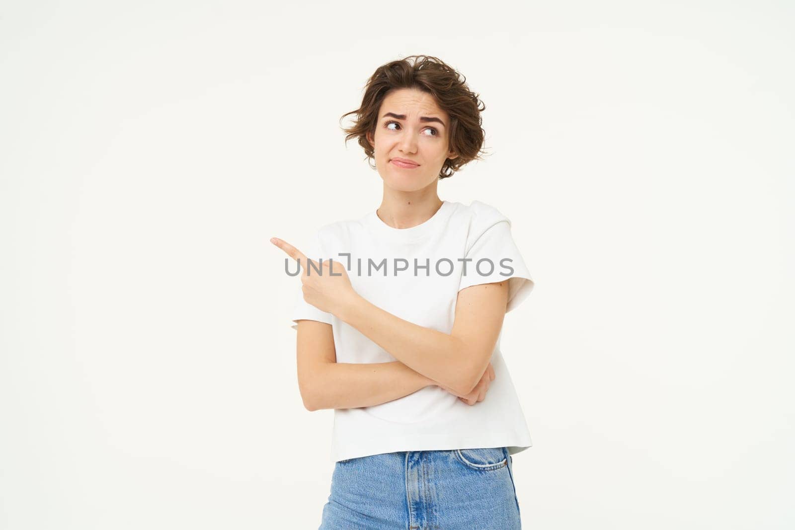 Image of woman with sceptical expression, pointing finger left, showing advertisement with doubtful face expression, standing over white studio background.