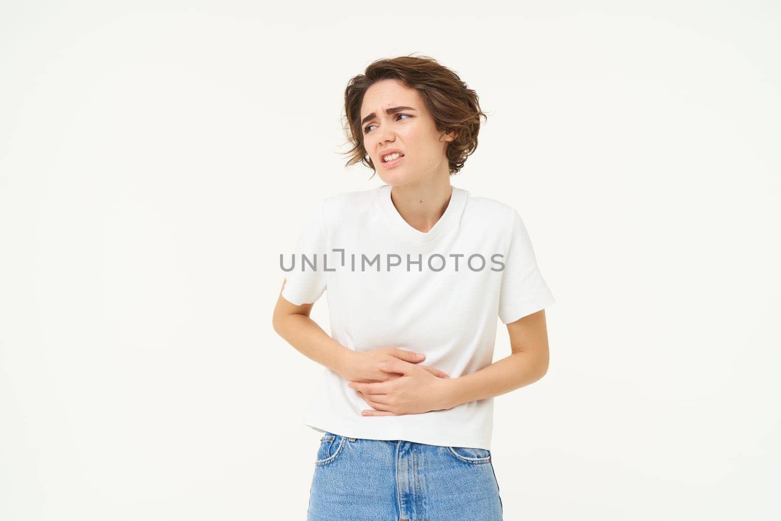 Woman grimacing from pain in stomach, stomachache, touches her belly, has painful periods, menstrual cramps, asks for painkillers, stands over white background.
