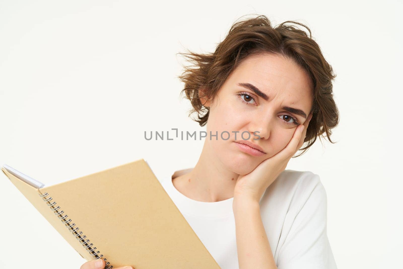 Image of troubled, upset young woman, student looks complicated, holds reading material homework, frowning with stressed face expression, standing over white background.