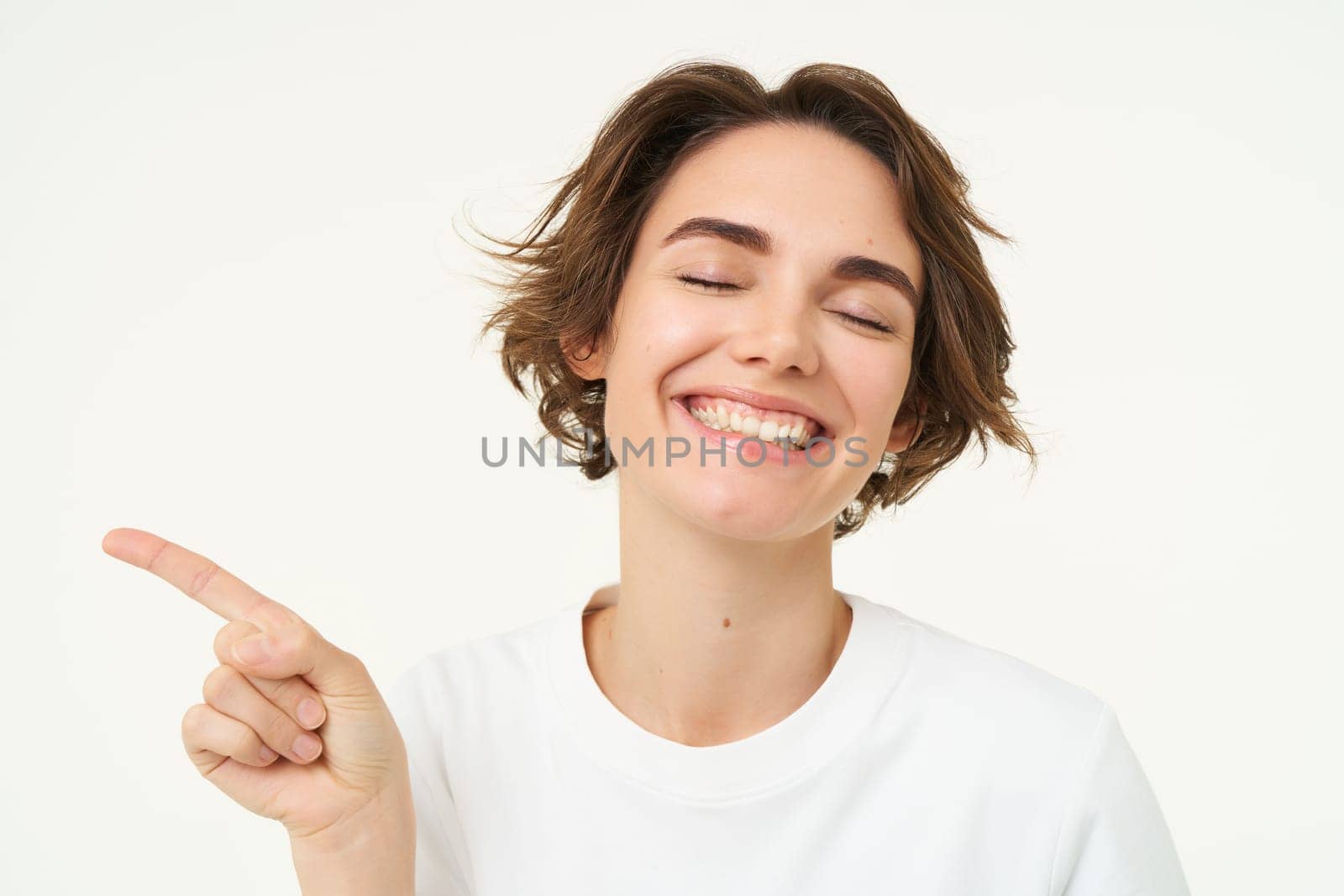 Portrait of candid young woman with short curly hair, pointing finger left at banner, showing product advertisement and smiling, posing over white background.
