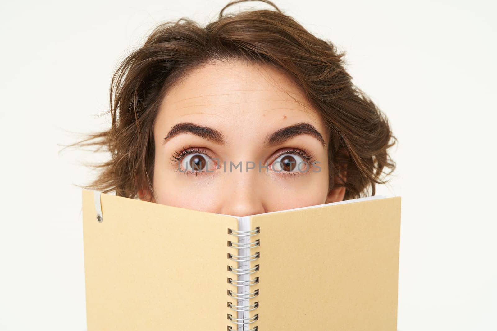 Close up portrait of girl face, hiding behind notebook, looking aside, standing over white background by Benzoix