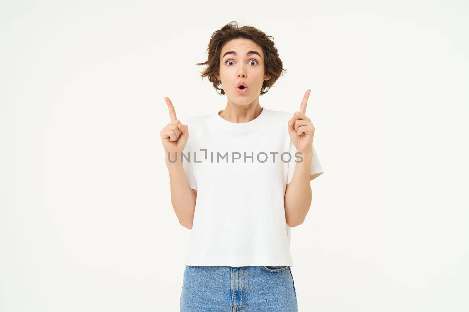 Image of brunette woman pointing fingers up, showing advertisement, gasping and looking amazed, saying wow, impressed by offer, standing over white background by Benzoix