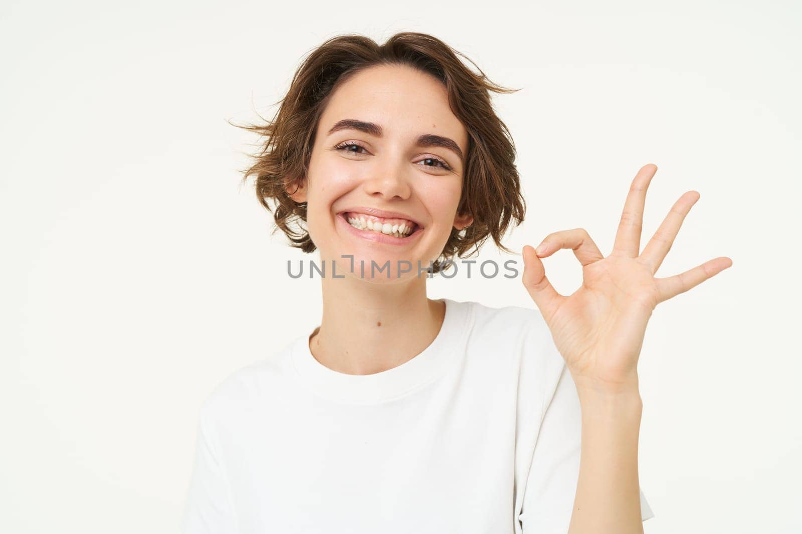 Close up portrait of satisfied, smiling young woman, shows okay, ok gesture, recommends something, gives positive feedback, says yes, white background.