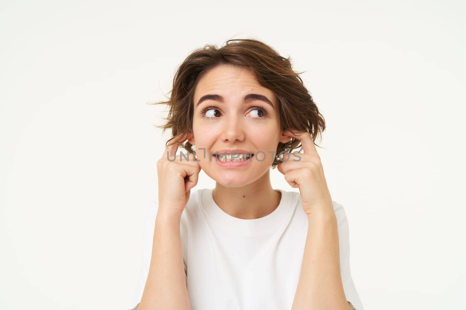 Close up portrait of woman smiling with awkward face expression, shuts her ears, doesnt want to listen, disturbed by loud noise, stands over white studio background by Benzoix