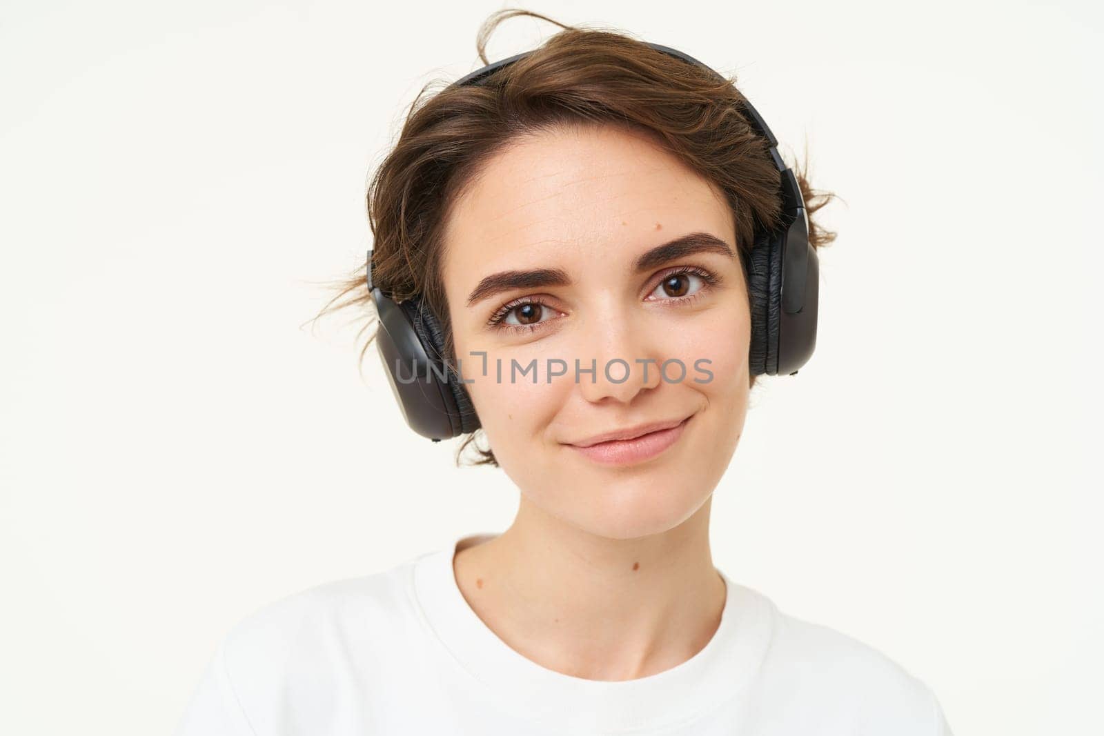 Portrait of young candid woman in wireless headphones, smiling, listening music in earphones, standing over white background.