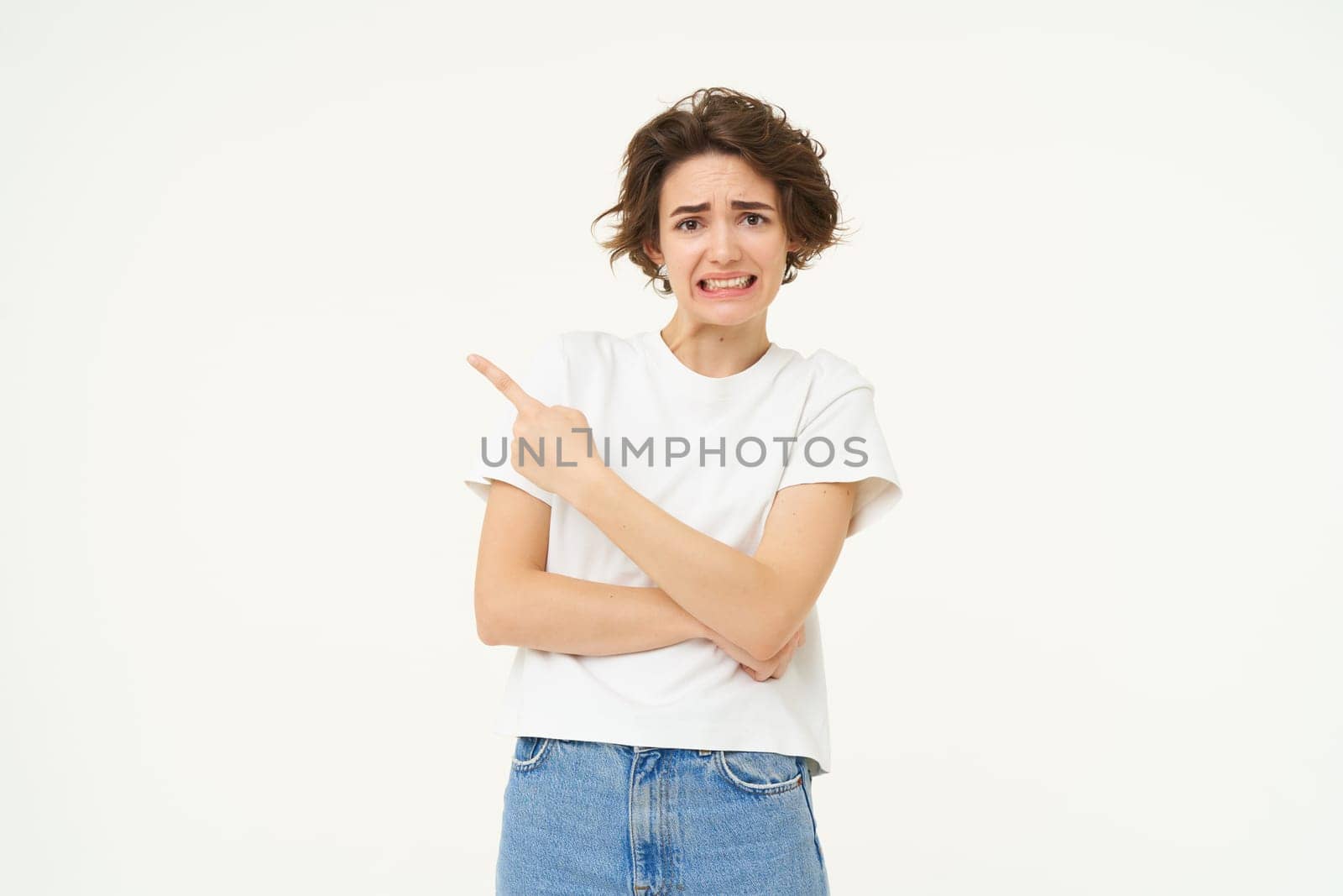 Portrait of woman feels discomfort, points finger left, shows advertisement and looks concerned, stands over white studio background by Benzoix