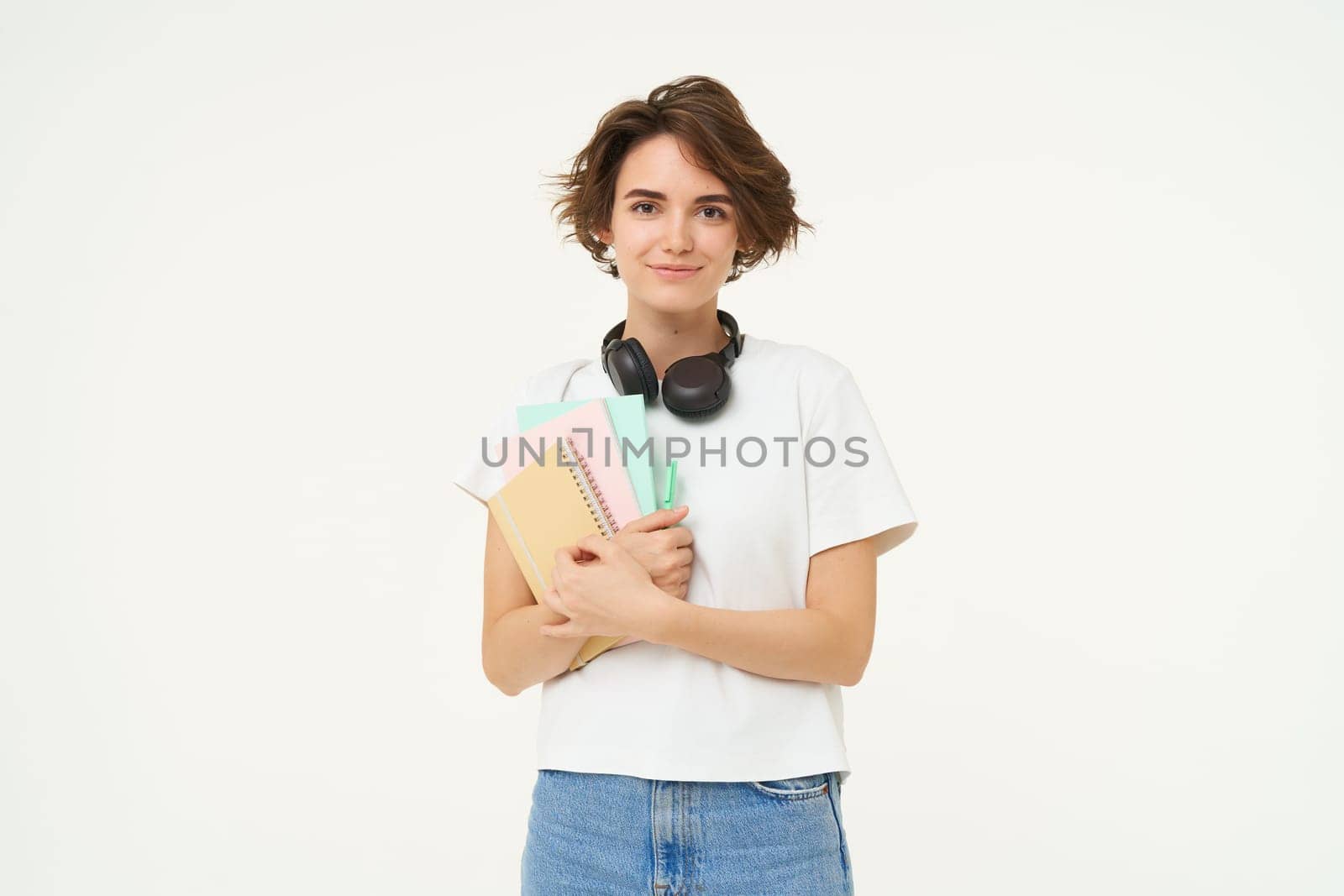 Image of brunette woman in headphones, standing with documents. Student with notes and workbooks posing against white background by Benzoix