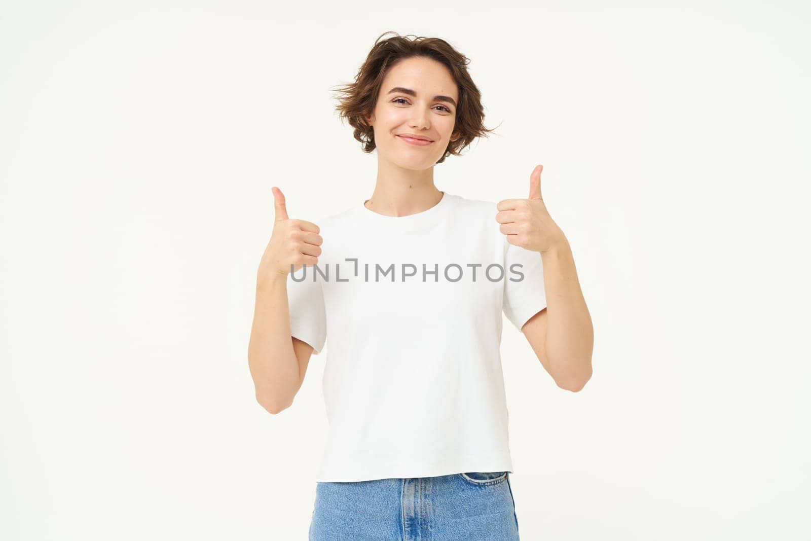 Image of young woman giving positive feedback, shows thumbs up and smiling, looks with confidence, stands over white background by Benzoix