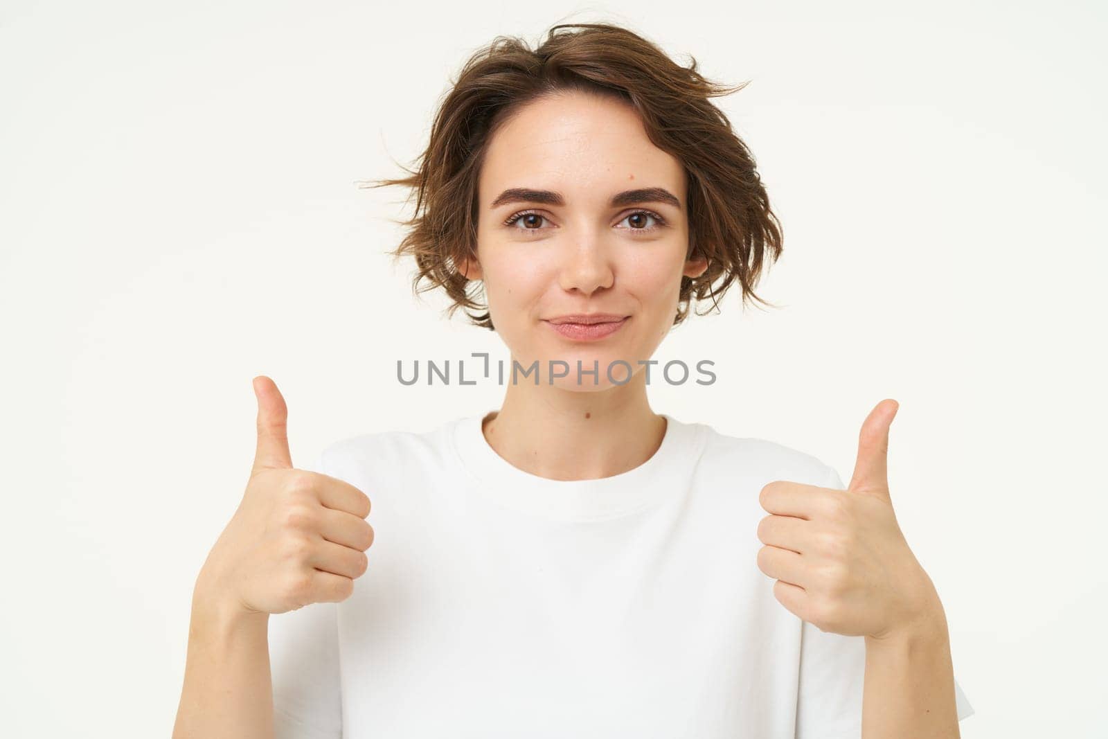 Close up of happy brunette woman, shows thumbs up, approves something, recommends, gives positive feedback, stands over white background.