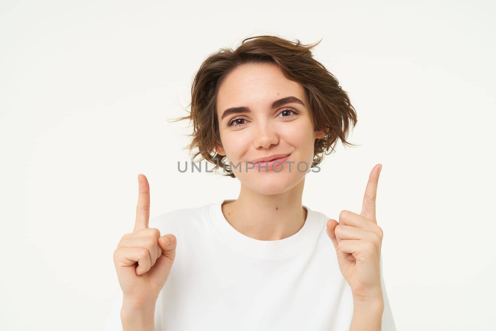 Portrait of cheerful girl with smiling face, pointing fingers up, showing banner or advertisement, look at top gesture, standing against white background.