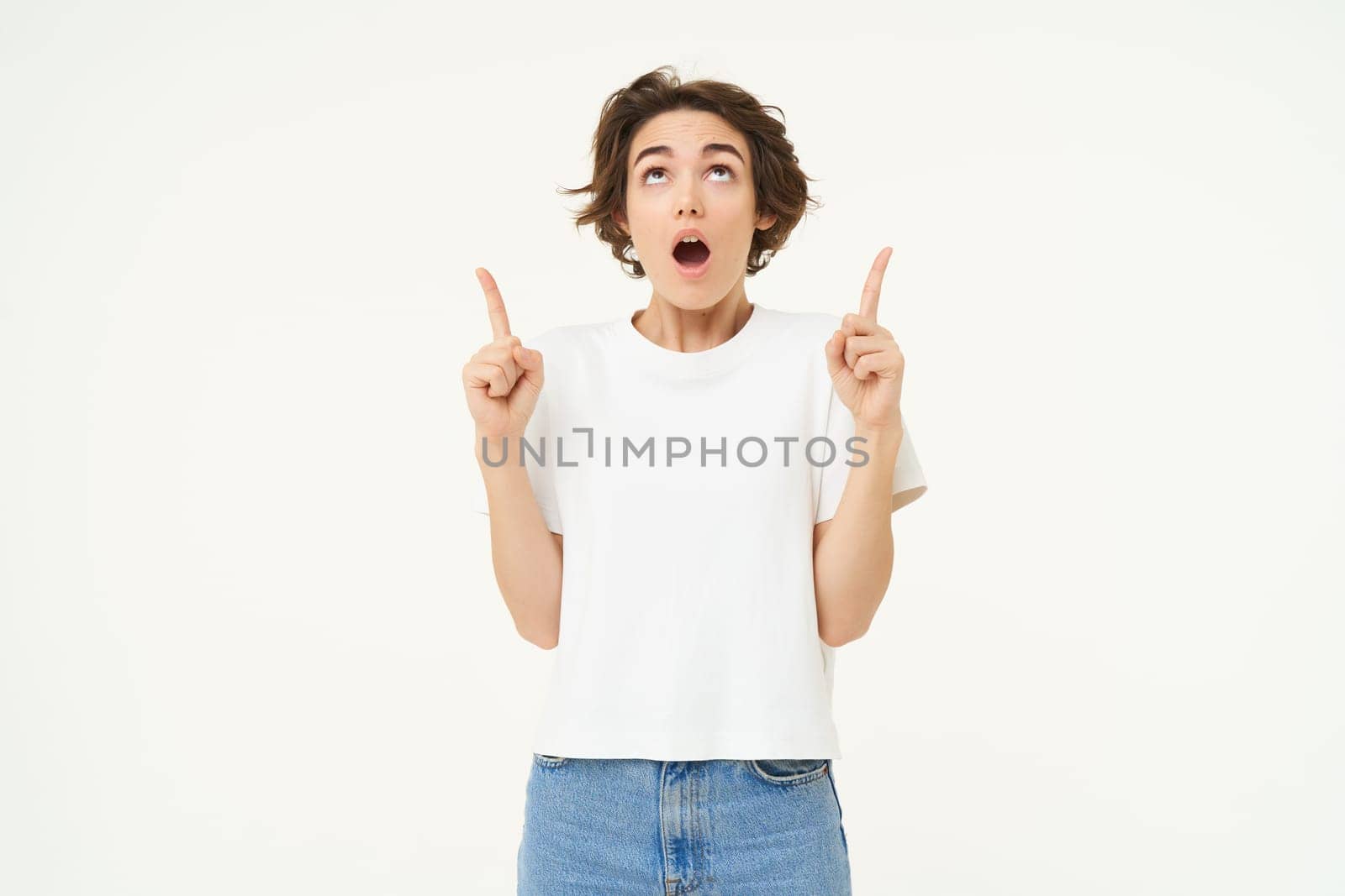 Portrait of woman with surprised face, amazed by promo offer, pointing fingers up and looking on top, standing against white background.