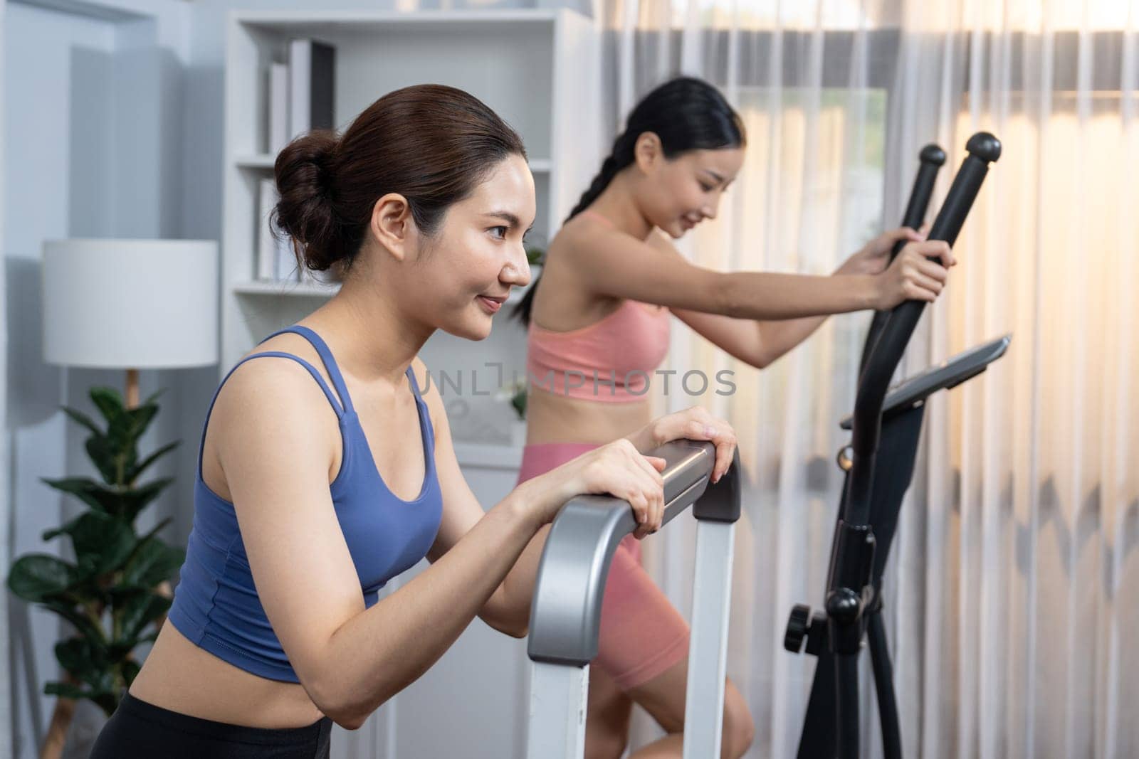 Energetic and strong athletic asian woman running on elliptical running machine at home with workout buddy or trainer. Pursuit of fit physique and commitment to healthy lifestyle. Vigorous
