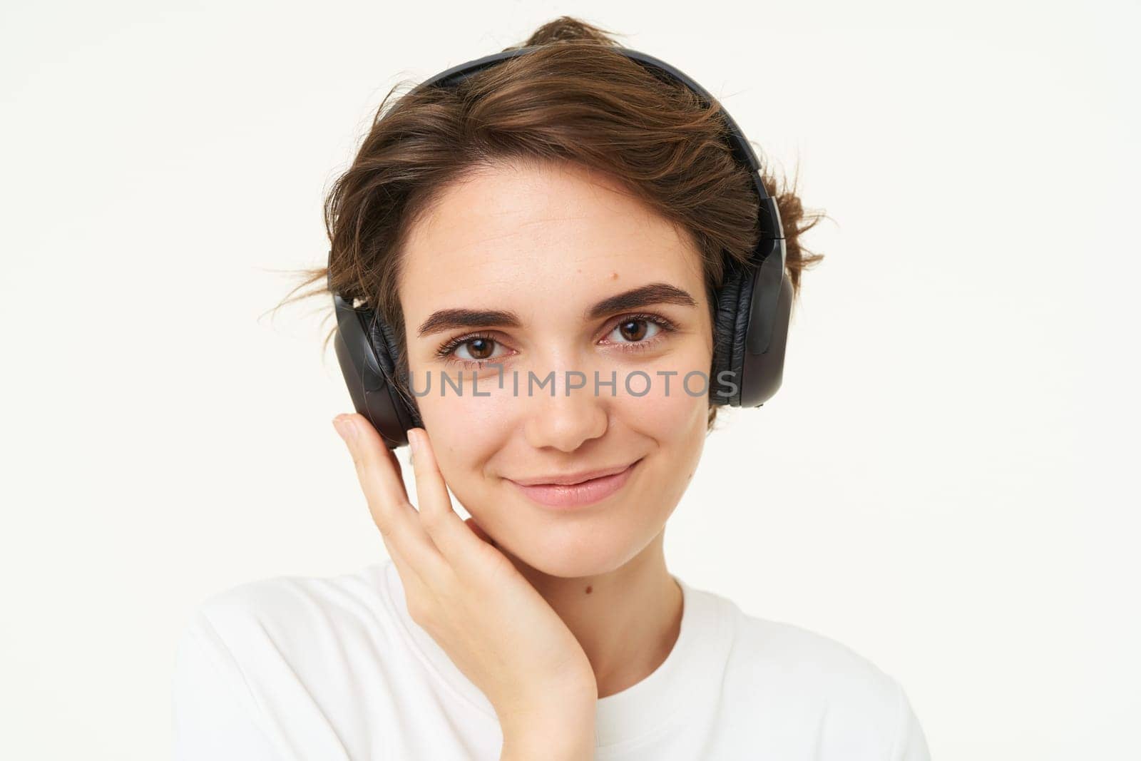 Image of young modern woman in wireless headphones, using earphones to listen to music, standing over white background. Copy space