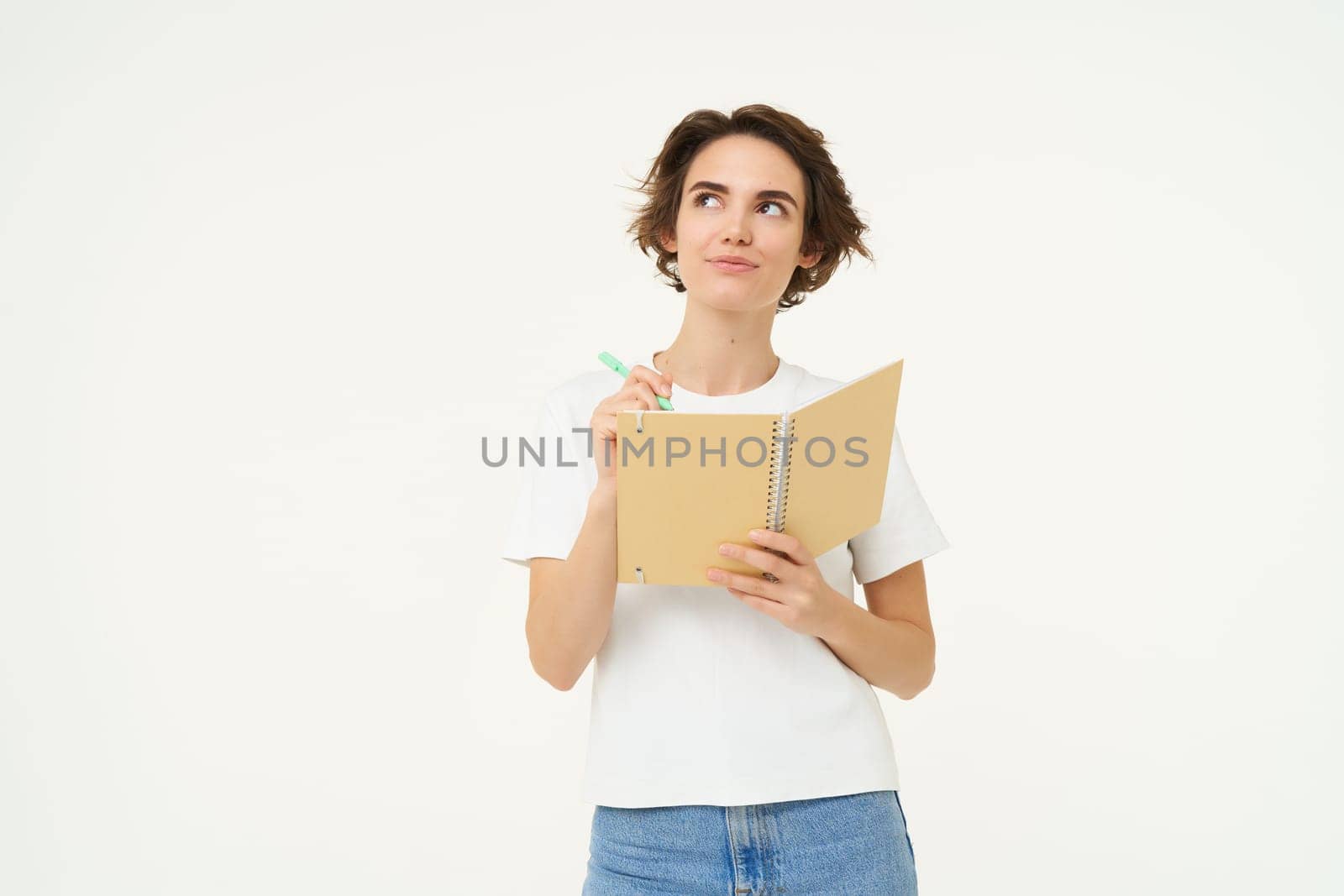 Portrait of creative girl, student doing homework, writing in notebook, standing with her diary, posing over white background.