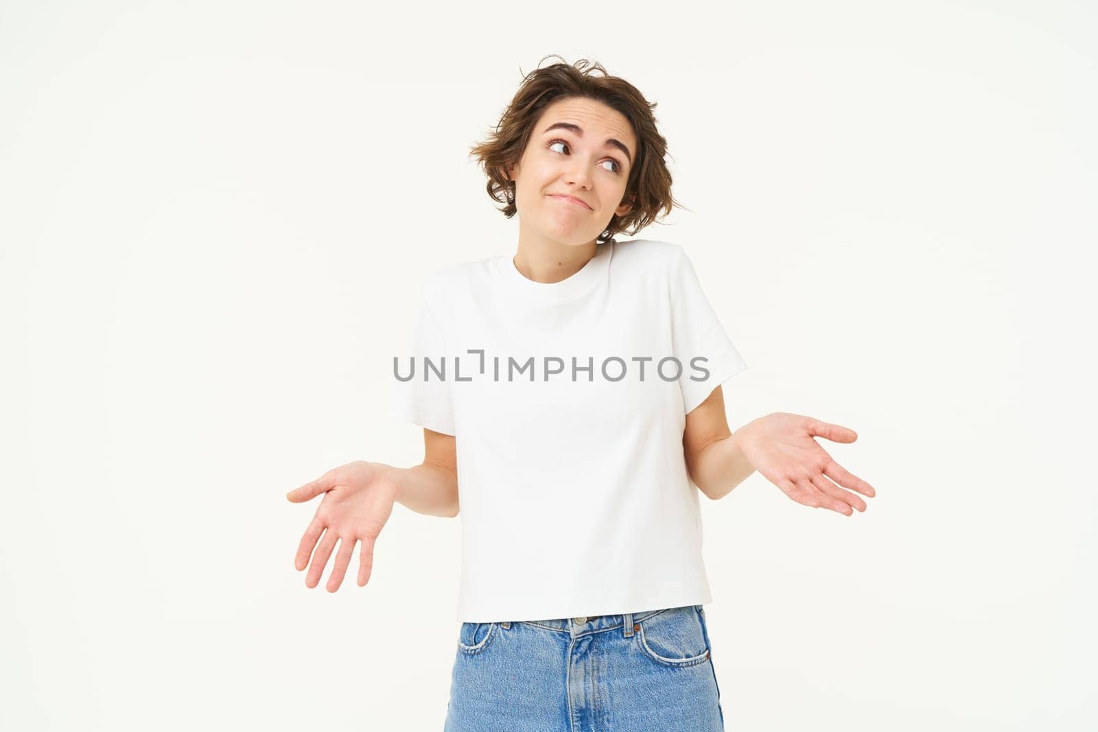 Portrait of woman spreads hands sideways and shrugs shoulders, looks clueless, dont know, stands unaware against white background.