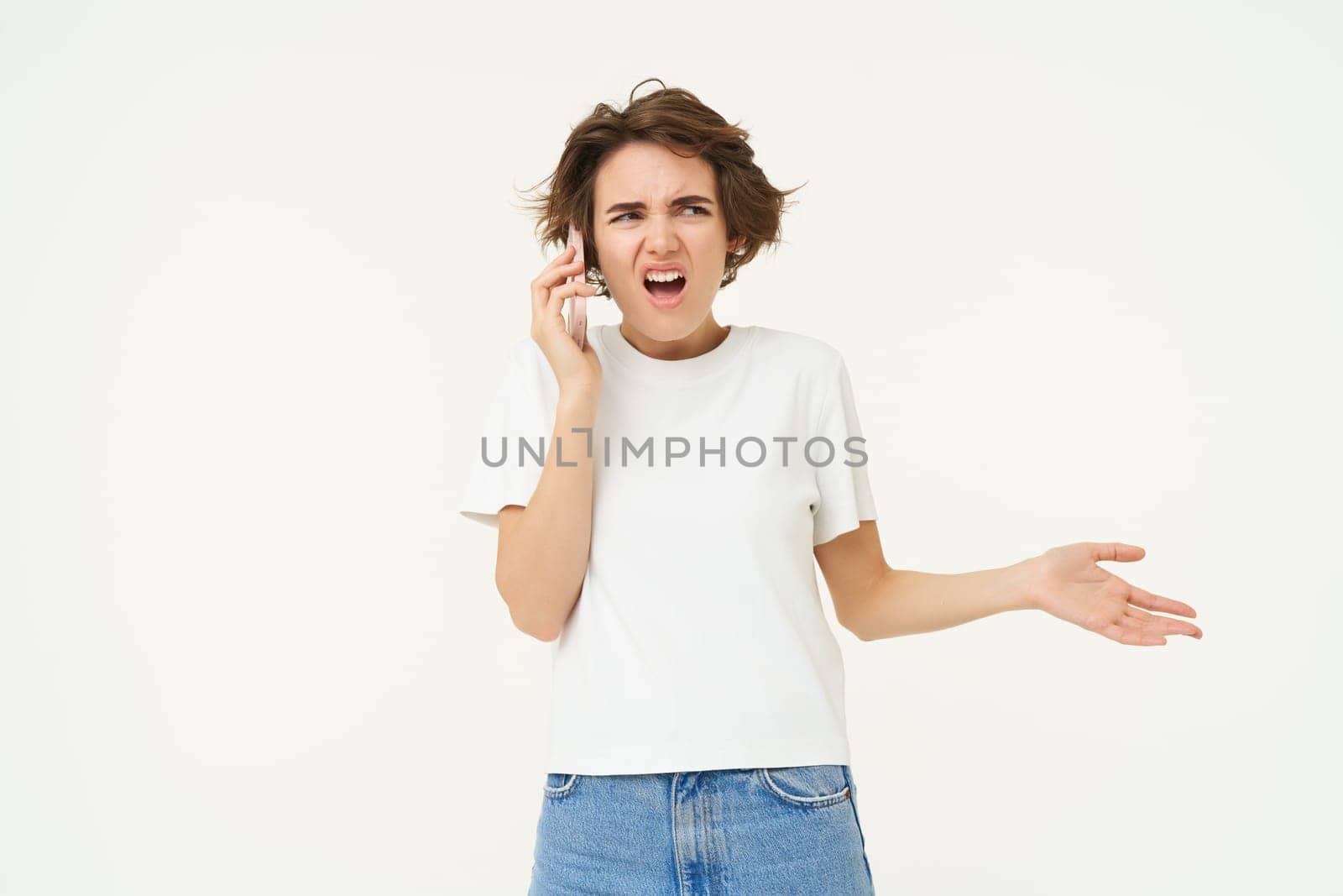 Portrait of shocked, angry woman talks on mobile phone with disappointed, furious face expression, standing over white background by Benzoix