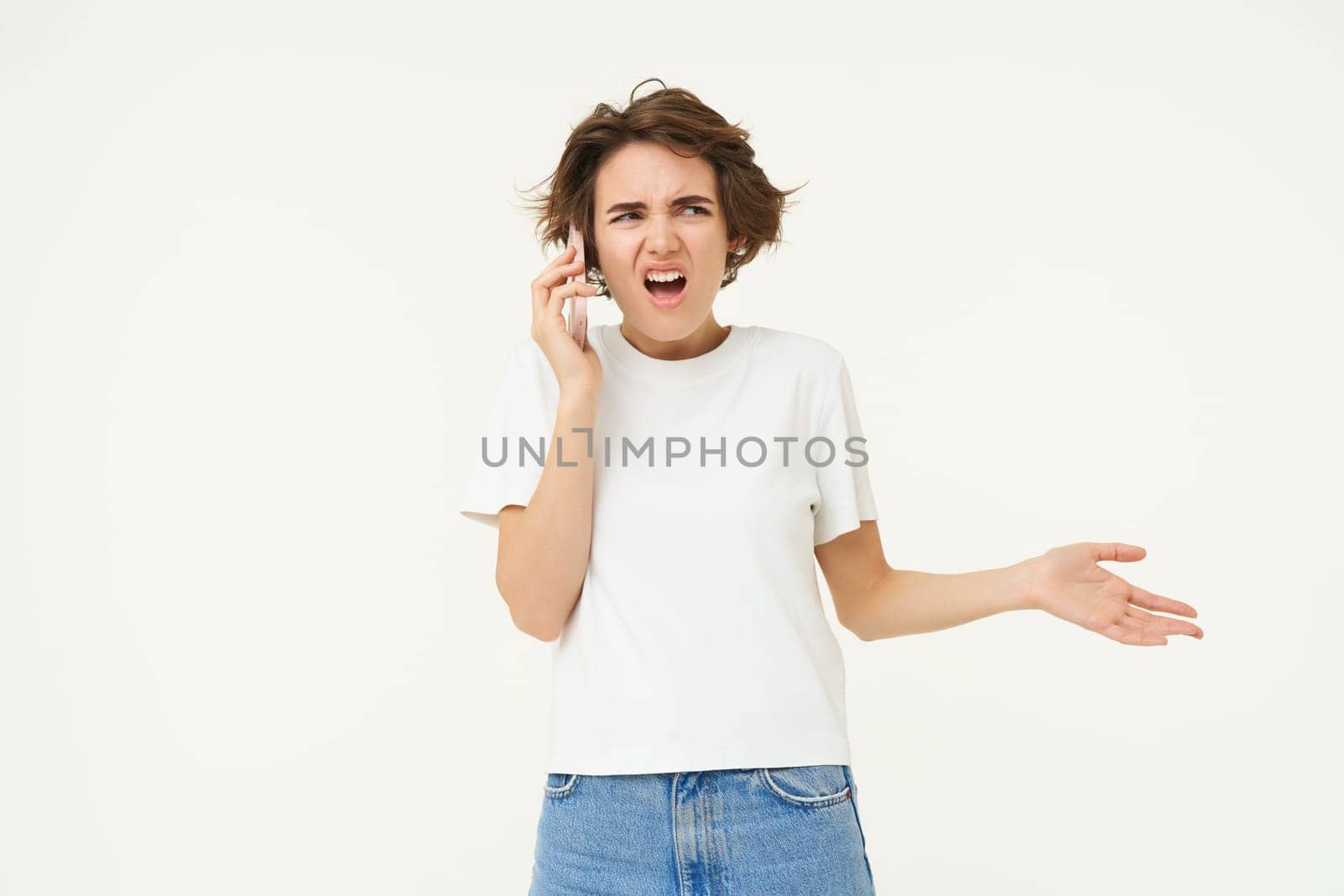 Portrait of shocked, angry woman talks on mobile phone with disappointed, furious face expression, standing over white background.