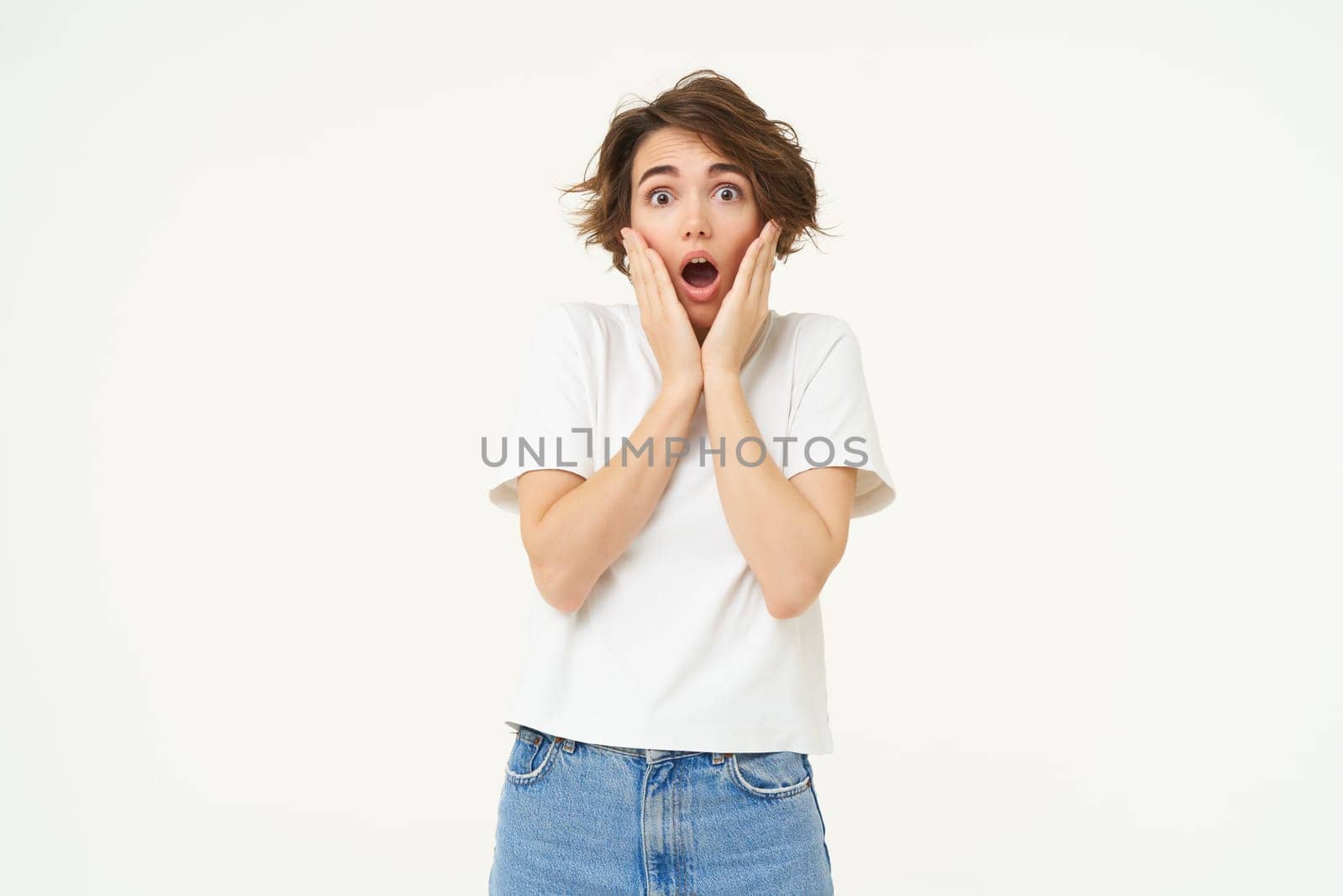 Portrait of girl with shocked, scared face, screams, holds hands on cheeks, stands over white studio background.