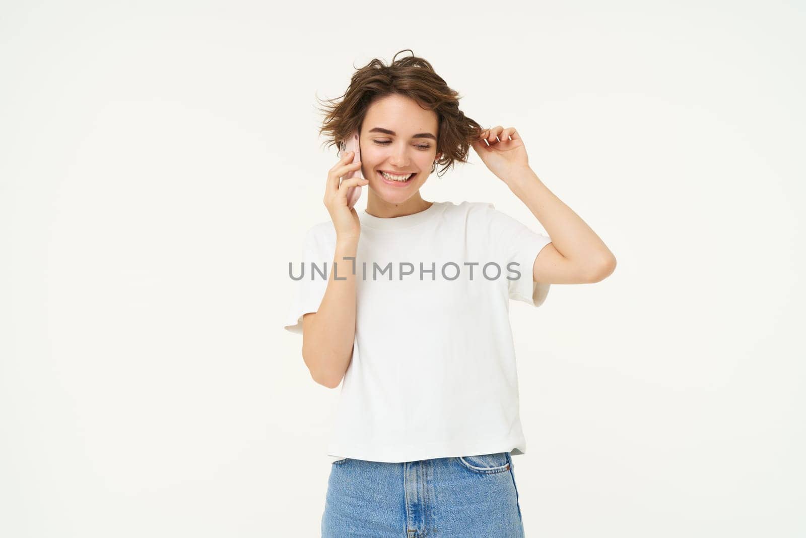 Cute and coquettish young woman calling someone, answer telephone, talking on smartphone and playing with her hair, standing over white background.