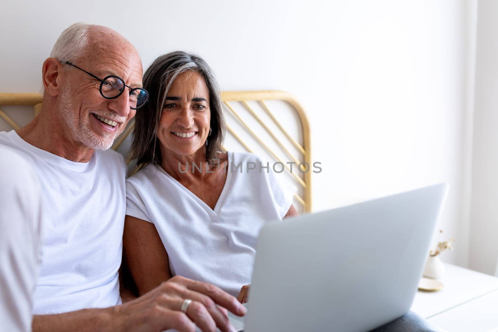 Mature couple using laptop together relaxing on bed. Copy space. Lifestyle and technology concepts.