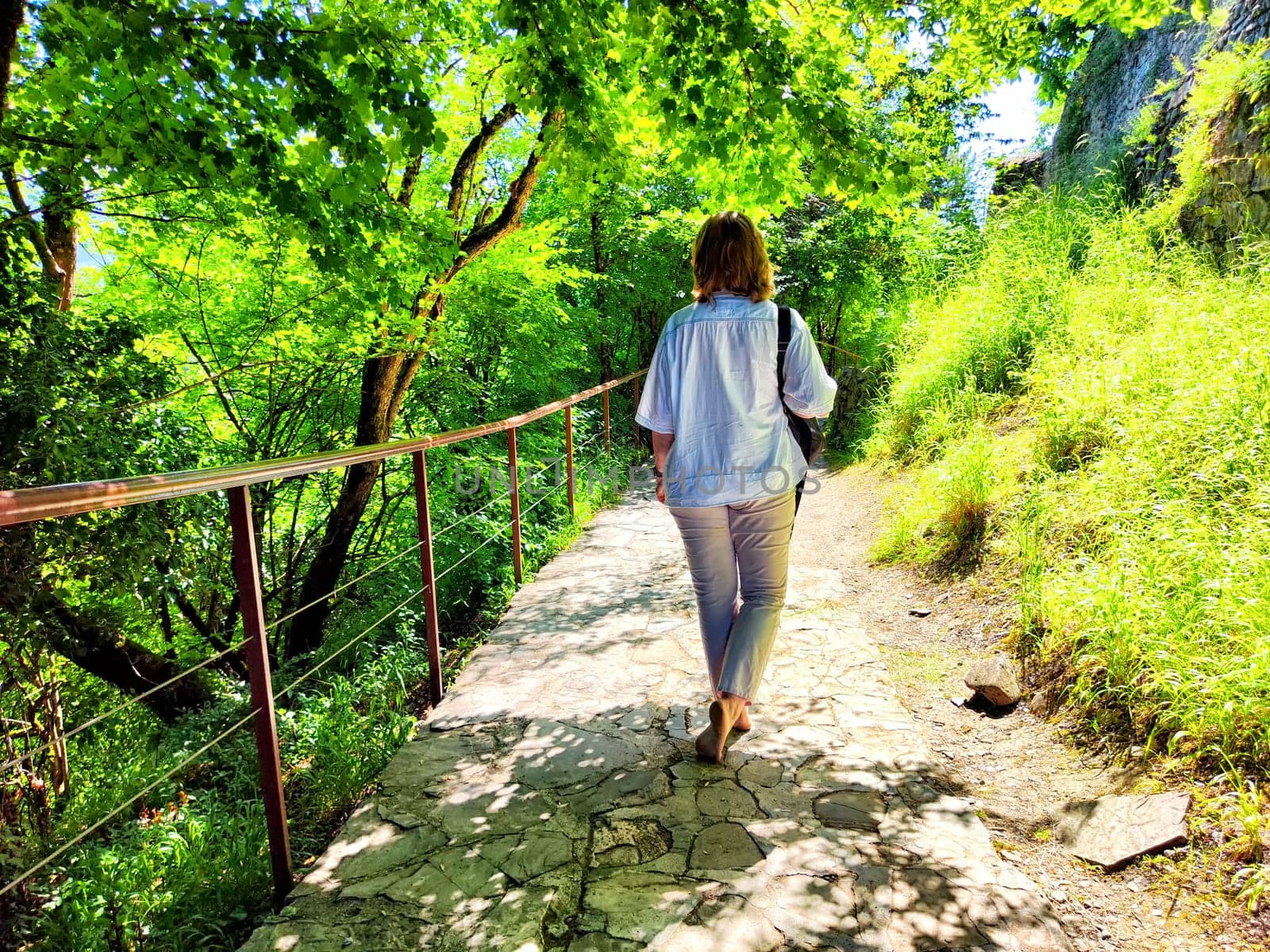 A woman strolls peacefully down a stone pathway enveloped by vibrant green foliage on a sunny day.