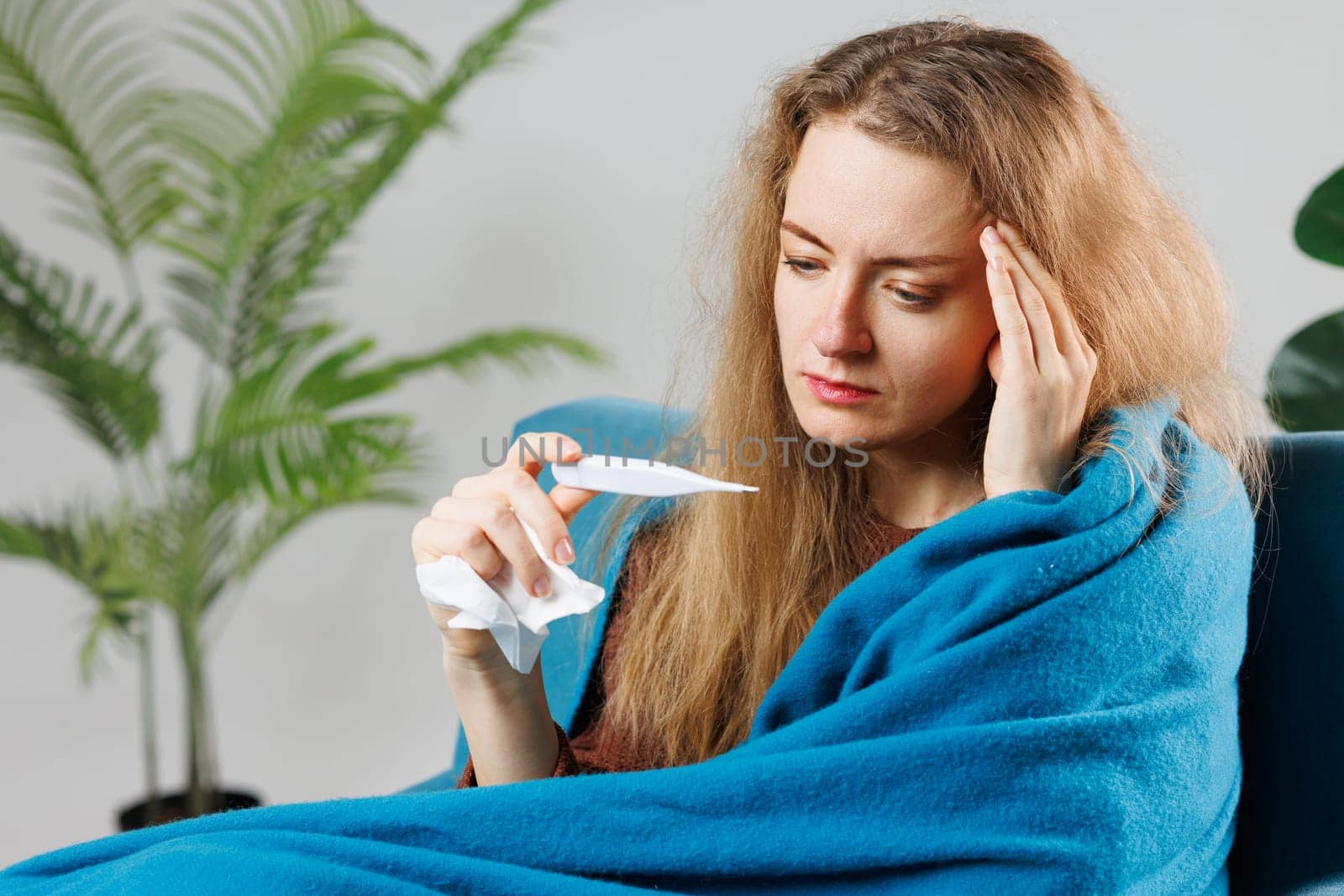 a sick girl with a cold or flu wrapped in a blue blanket measures her temperature at home, symptoms, treatment of viral diseases
